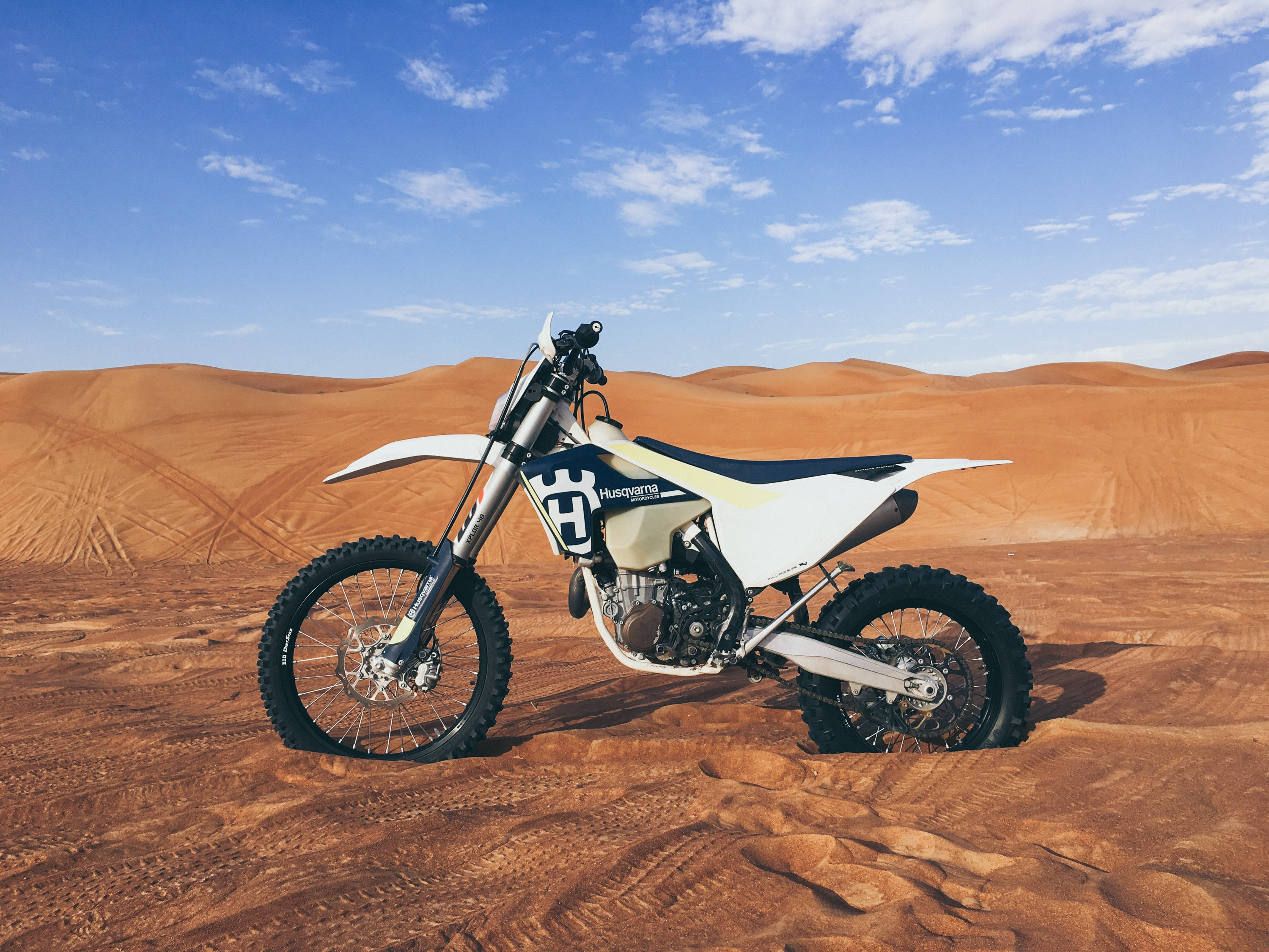 機車 桌布 壁紙 — 一輛白色 Husqvarna 越野摩托車矗立在金色沙漠沙丘上，天空湛藍，白雲點綴。