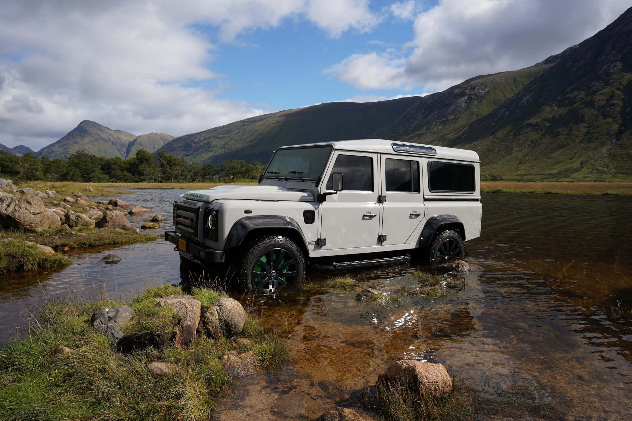 Land Rover Defender 桌布 壁紙 — 一輛白色 Land Rover Defender 在部分多雲的天空下，涉水穿過一條被岩石地形和遠處綠色山丘包圍的淺山溪。
