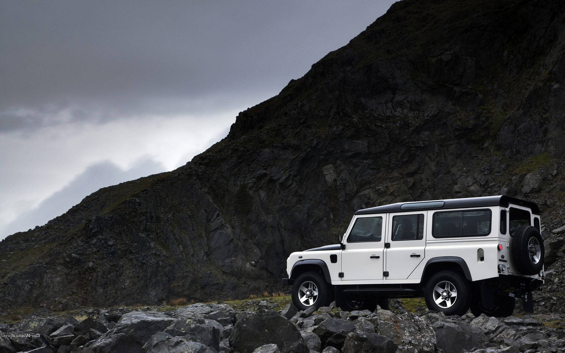 荒原路華 桌布 壁紙 — 一輛白色 Land Rover Defender 在陰沉多雲的天空下，在陡峭的黑暗山脈前的岩石地形上堅固地站立。