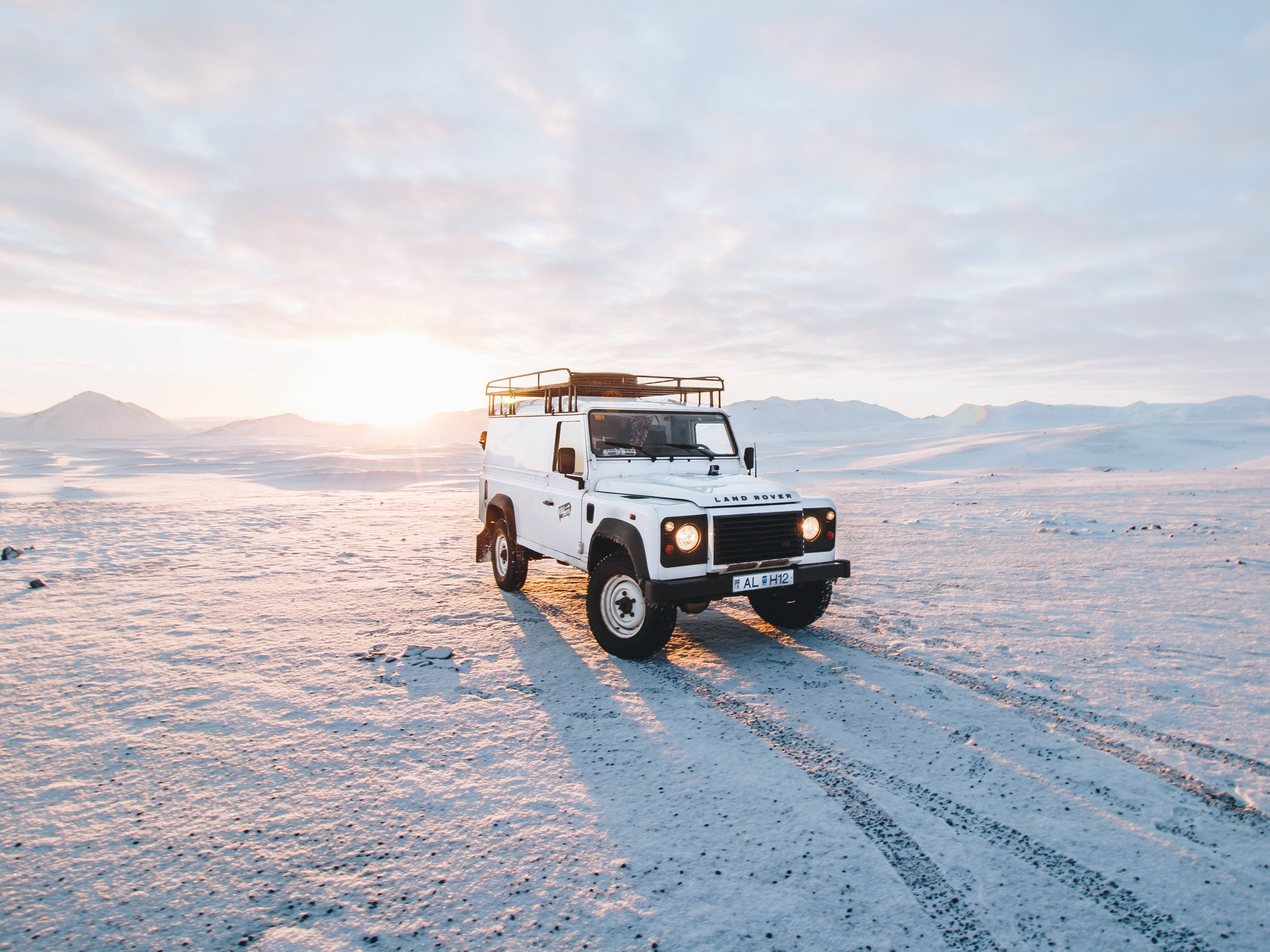 荒原路華 桌布 壁紙 — 一輛白色 Land Rover Defender 在日出時分駛過雪覆蓋的沙漠，遠山在柔和粉彩天空下。