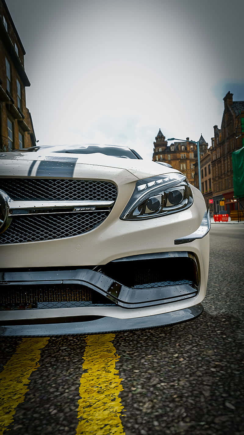 電玩遊戲 桌布 壁紙 — 一輛白色 Mercedes-AMG 跑車停泊在濕滑的城市街道上，背景有黃色道路標線和歷史建築的低角度拍攝。