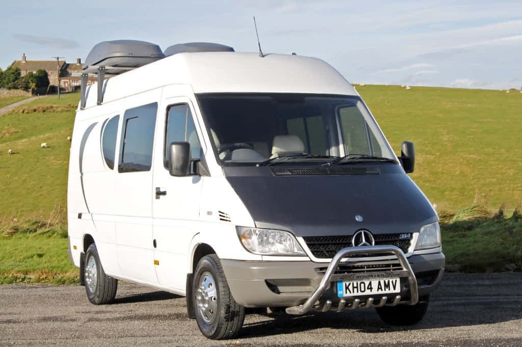 Mercedes Benz Sprinter 桌布 壁紙 — 一輛白色 Mercedes-Benz Sprinter 露營車，車頂裝有行李箱，停泊在碎石路上，旁邊是長滿草的山丘，天空局部多雲。