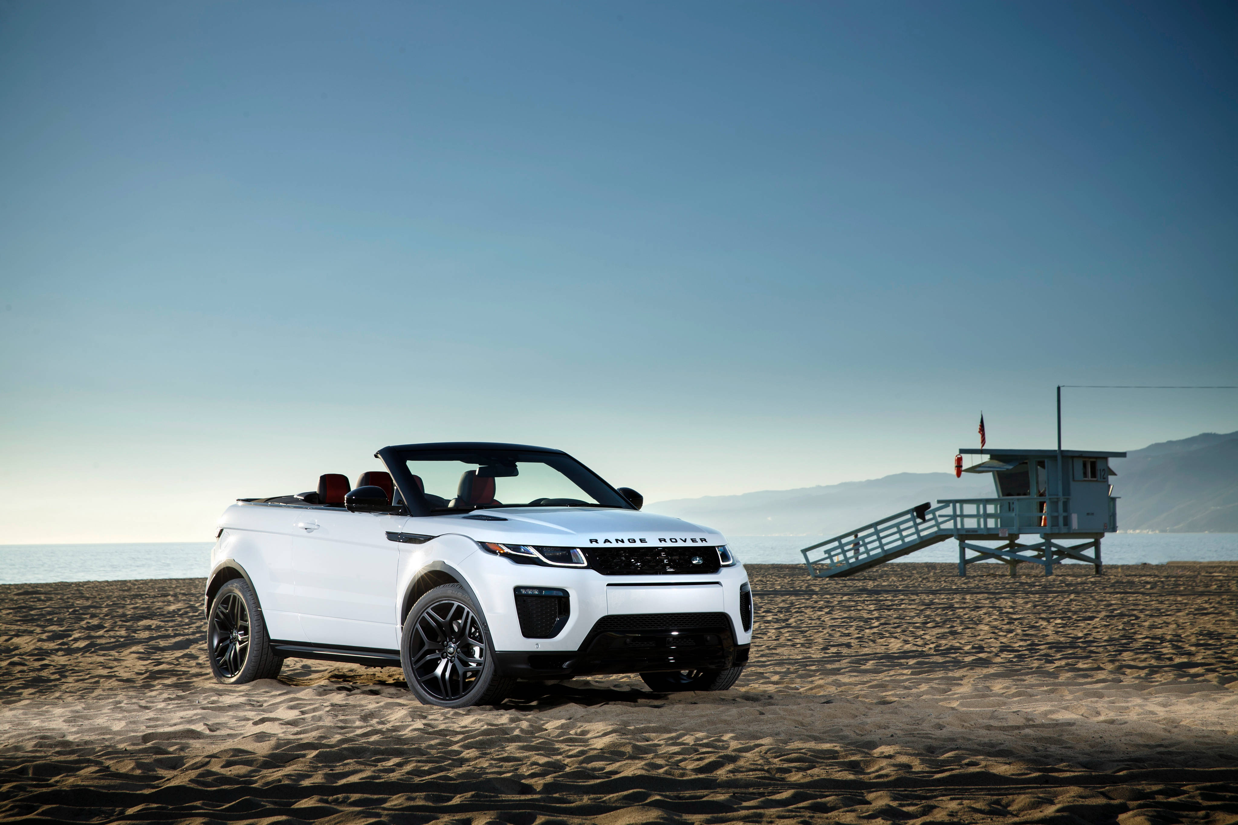 荒原路華 桌布 壁紙 — 一輛白色 Range Rover Evoque Convertible 停泊在沙灘上，旁邊有一個救生員瞭望塔，天空清澈湛藍。