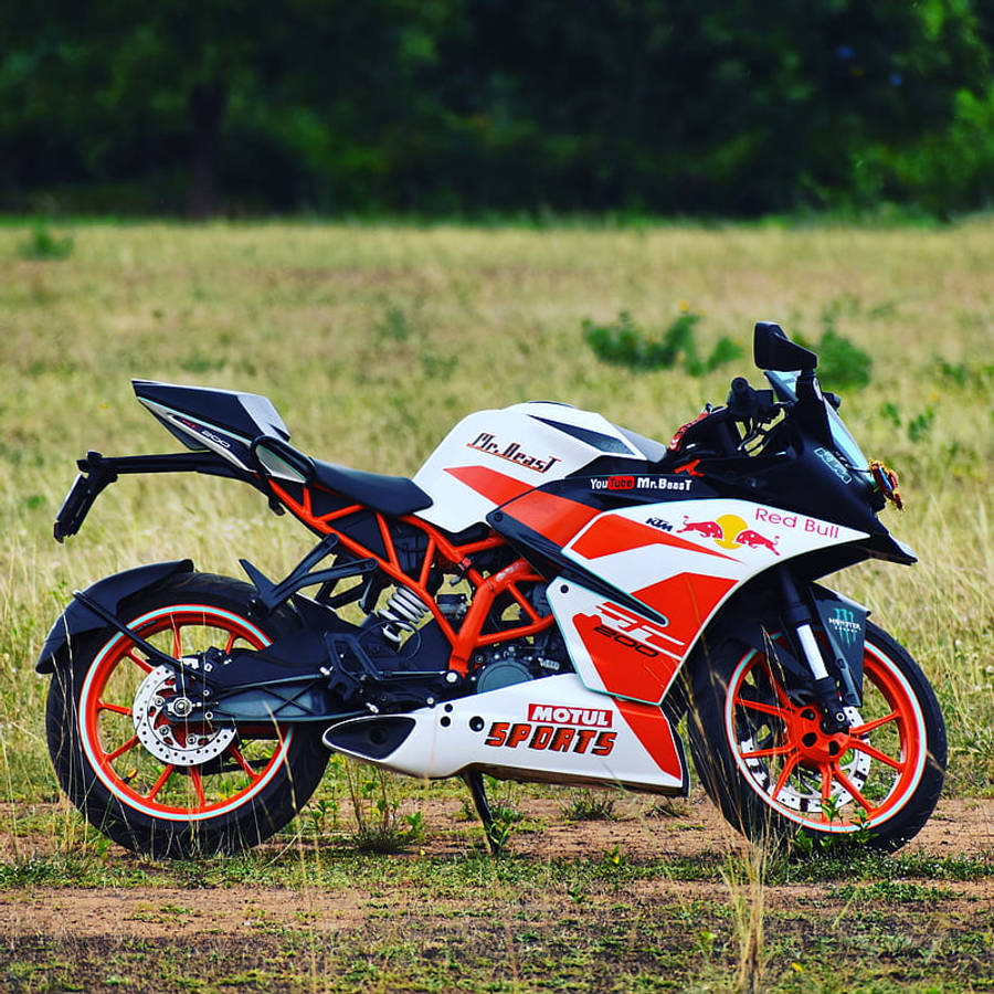 KTM RC 200 桌布 壁紙 — 一輛白色、橙色和黑色的KTM RC390摩托車，帶有Red Bull和MrBeast貼紙，停放在陽光明媚的草地上。