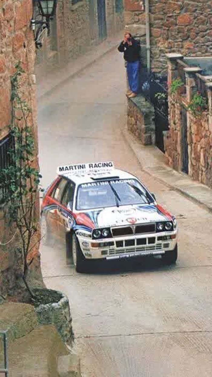 Lancia 桌布 壁紙 — 一輛白色、紅色和藍色的 Martini Racing Lancia 拉力賽車在狹窄的石頭村莊街道上疾馳，揚起灰塵，一名觀眾在牆上觀看。