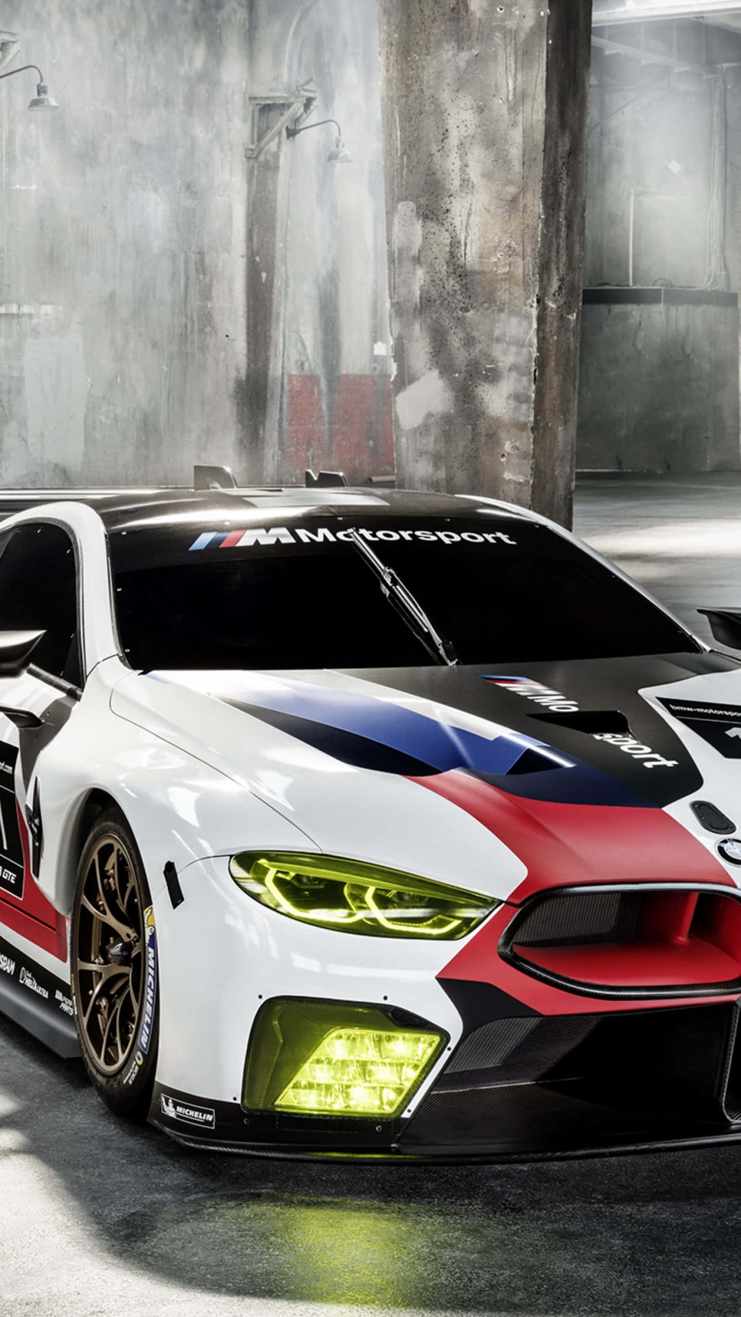 Bmw M8 4K 桌布 壁紙 — 一輛白色、紅色和黑色的BMW M8 GTLM賽車，帶有發光的黃色頭燈，停在粗糙的混凝土車庫中。