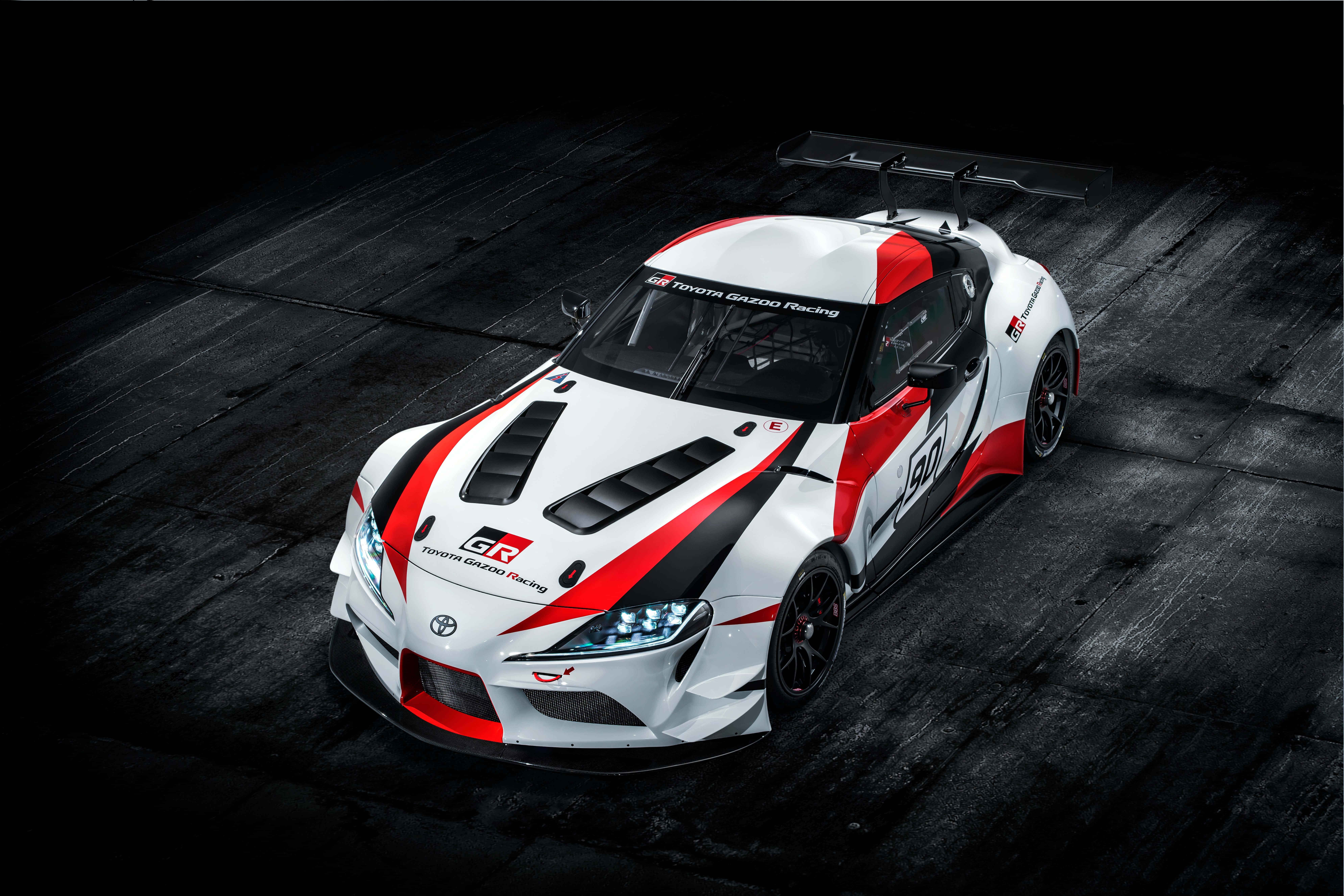 Supra 桌布 壁紙 — 一輛白色、紅色和黑色的豐田GR賽車，帶有大型後擾流板，在戲劇性的工作室燈光下放置在深色木地板上。