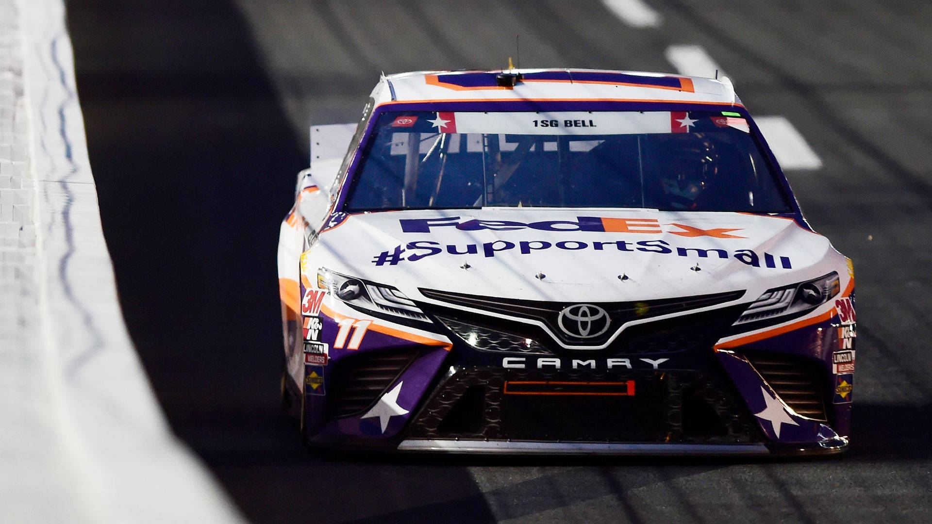 Denny Hamlin 桌布 壁紙 — 一輛白色、紫色和橙色的FedEx Toyota Camry賽車，帶有醒目的贊助商貼紙和星星圖案，在賽道上飛馳。