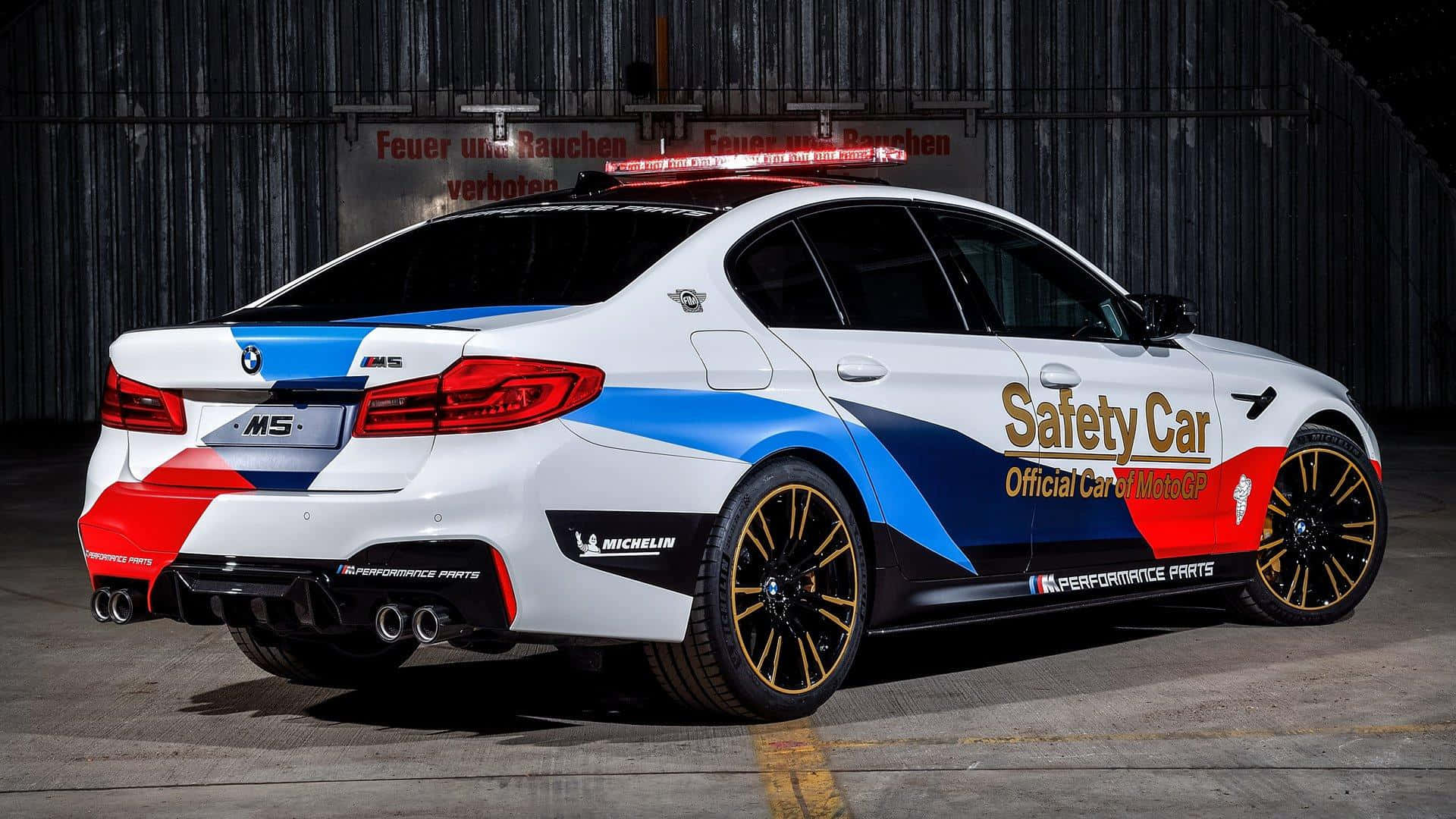 Safety Car 桌布 壁紙 — 一輛白色、藍色和紅色的BMW M5安全車，配備金色輪圈，停放在燈光昏暗的車庫中。