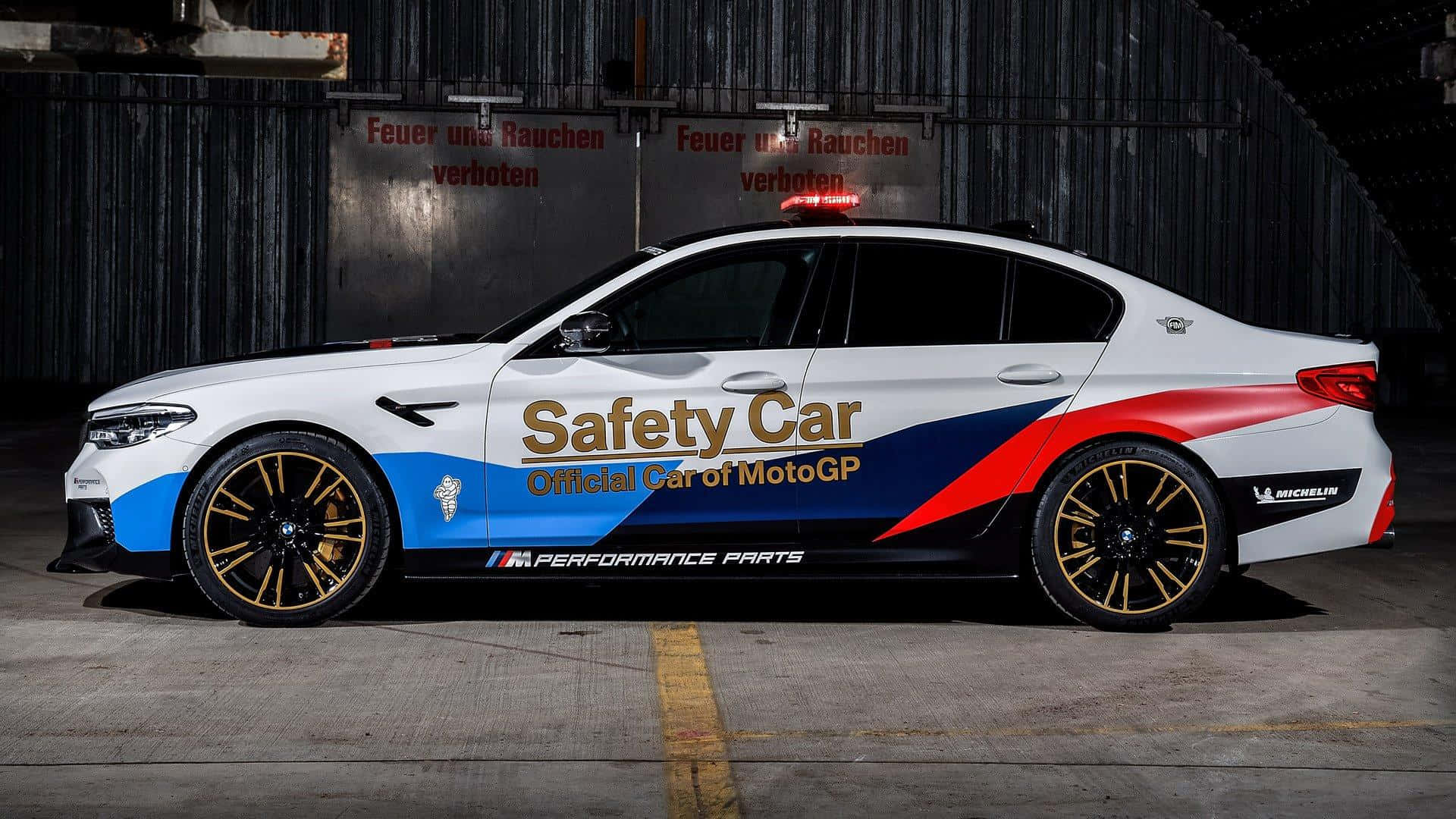 Safety Car 桌布 壁紙 — 一輛白色、藍色和紅色的BMW M5 MotoGP安全車停在一個光線昏暗的工業車庫內，帶有德文警告標誌。