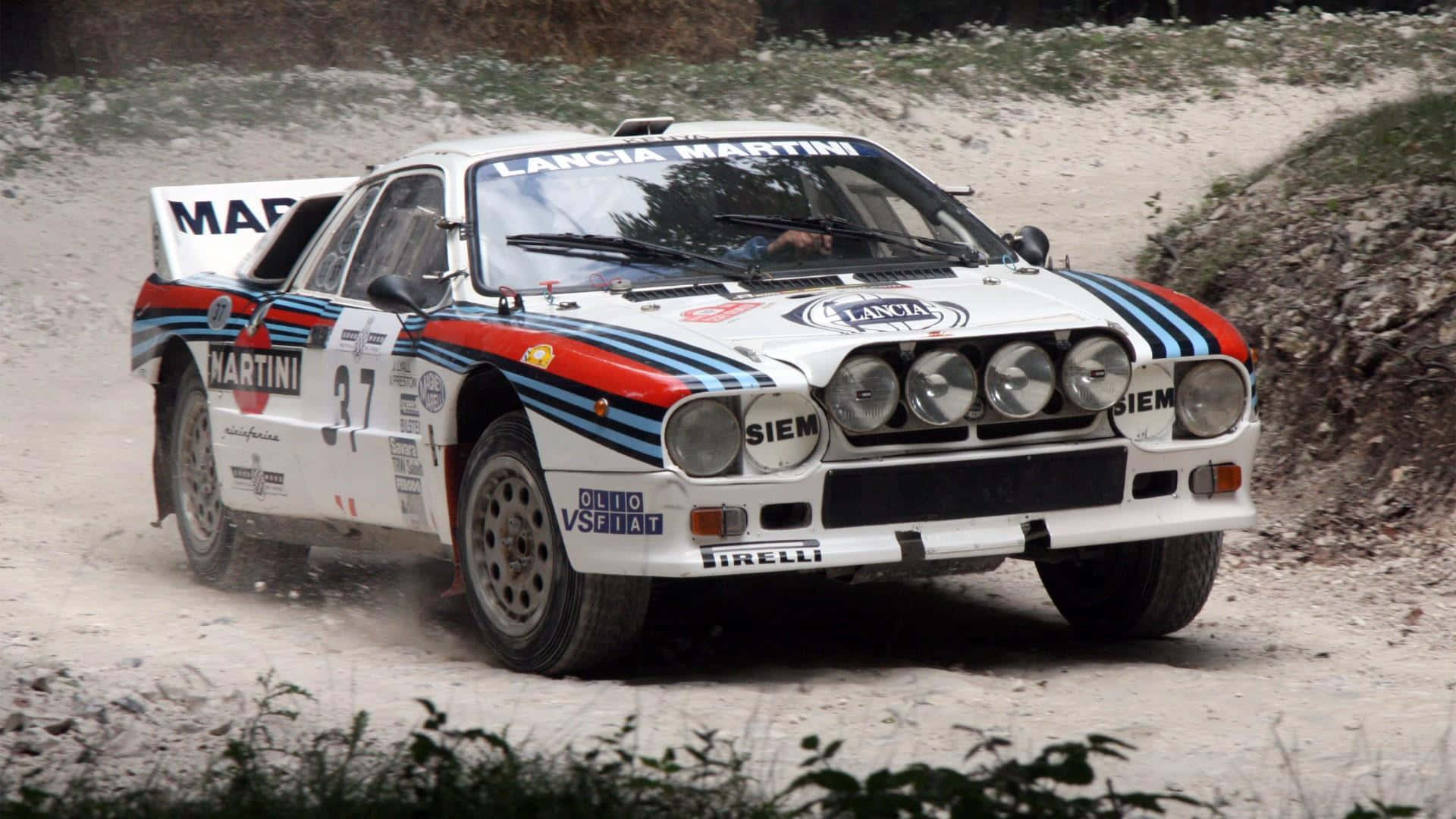 Lancia 桌布 壁紙 — 一輛白色、藍色和紅色的藍旗亞拉力賽車在崎嶇的泥路上疾馳，揚起灰塵。