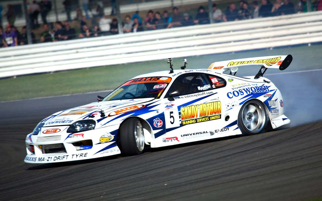 race car 桌布 壁紙 — 一輛白色、藍色和黃色的甩尾車，編號五，在賽道上燒胎冒煙，背景有觀眾。