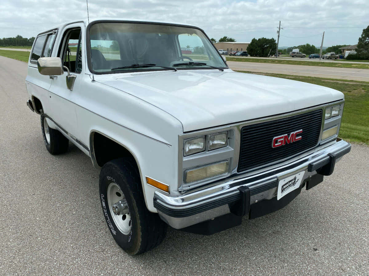 Gmc Jimmy 桌布 壁紙 — 一輛白色1980年代GMC Jimmy SUV停放在碎石地上，天空多雲，背景是郊區建築物。