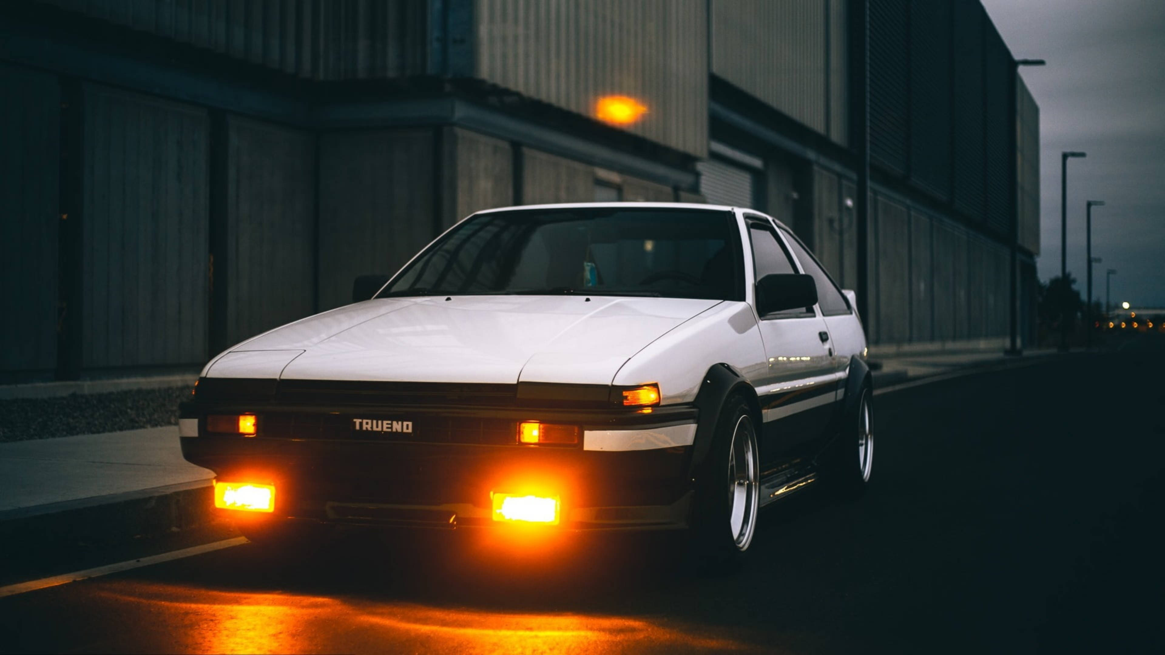 汽車 桌布 壁紙 — 一輛白色AE86 Trueno跑車，帶有發光的橙色頭燈，停泊在黃昏時潮濕的城市街道上。