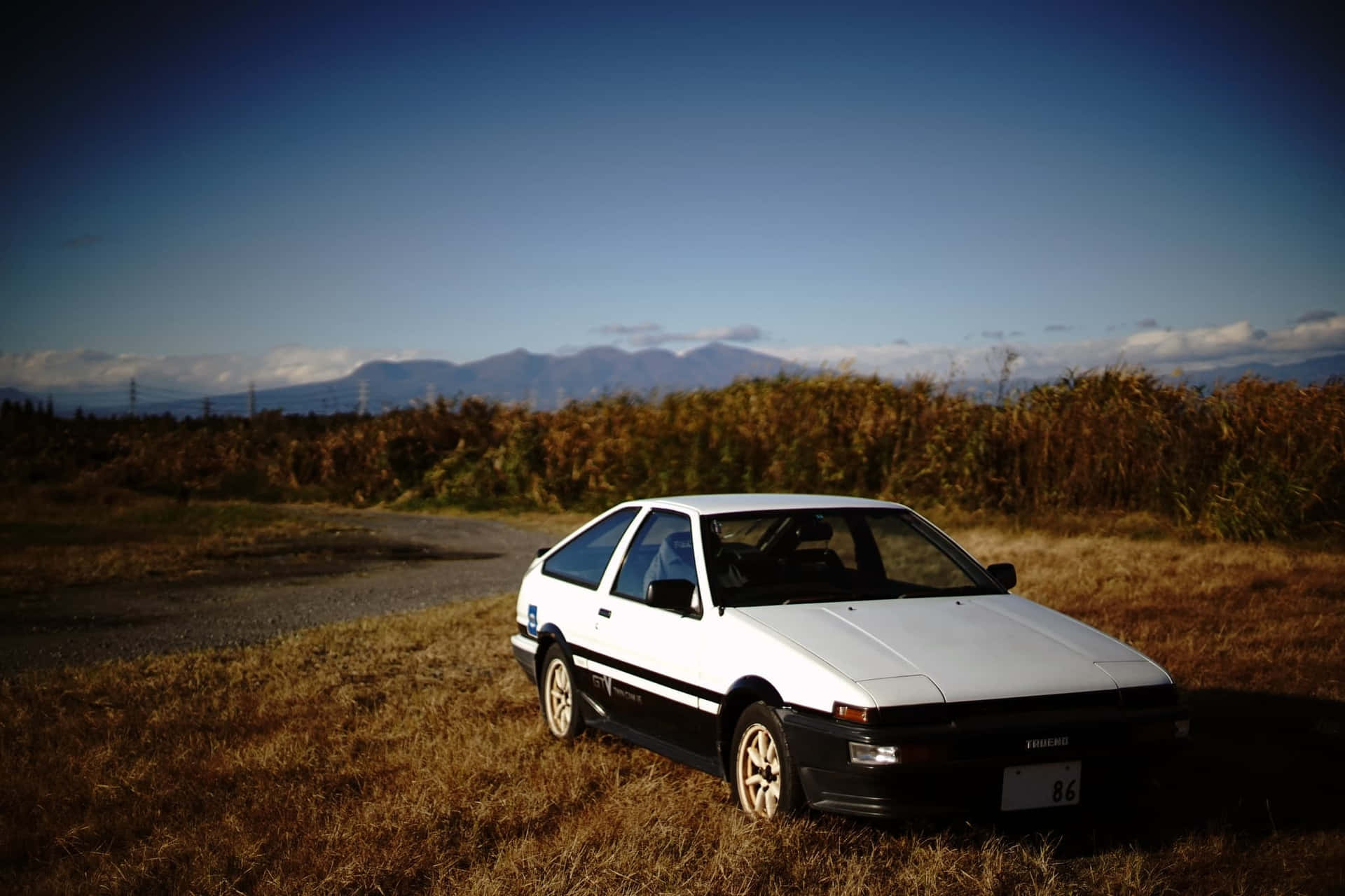 白色AE86跑車停泊在田野中，背景有山。 — Toyota Supra Mk4 壁紙