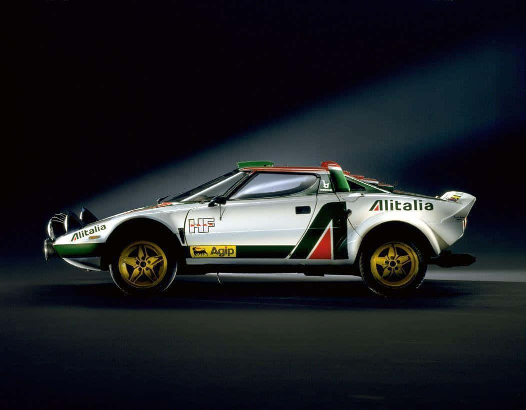 Lancia 桌布 壁紙 — 一輛白色Alitalia Lancia Delta S4拉力賽車，帶有綠色、紅色和黃色點綴，在黑暗的攝影棚背景下被聚光燈戲劇性地照亮。