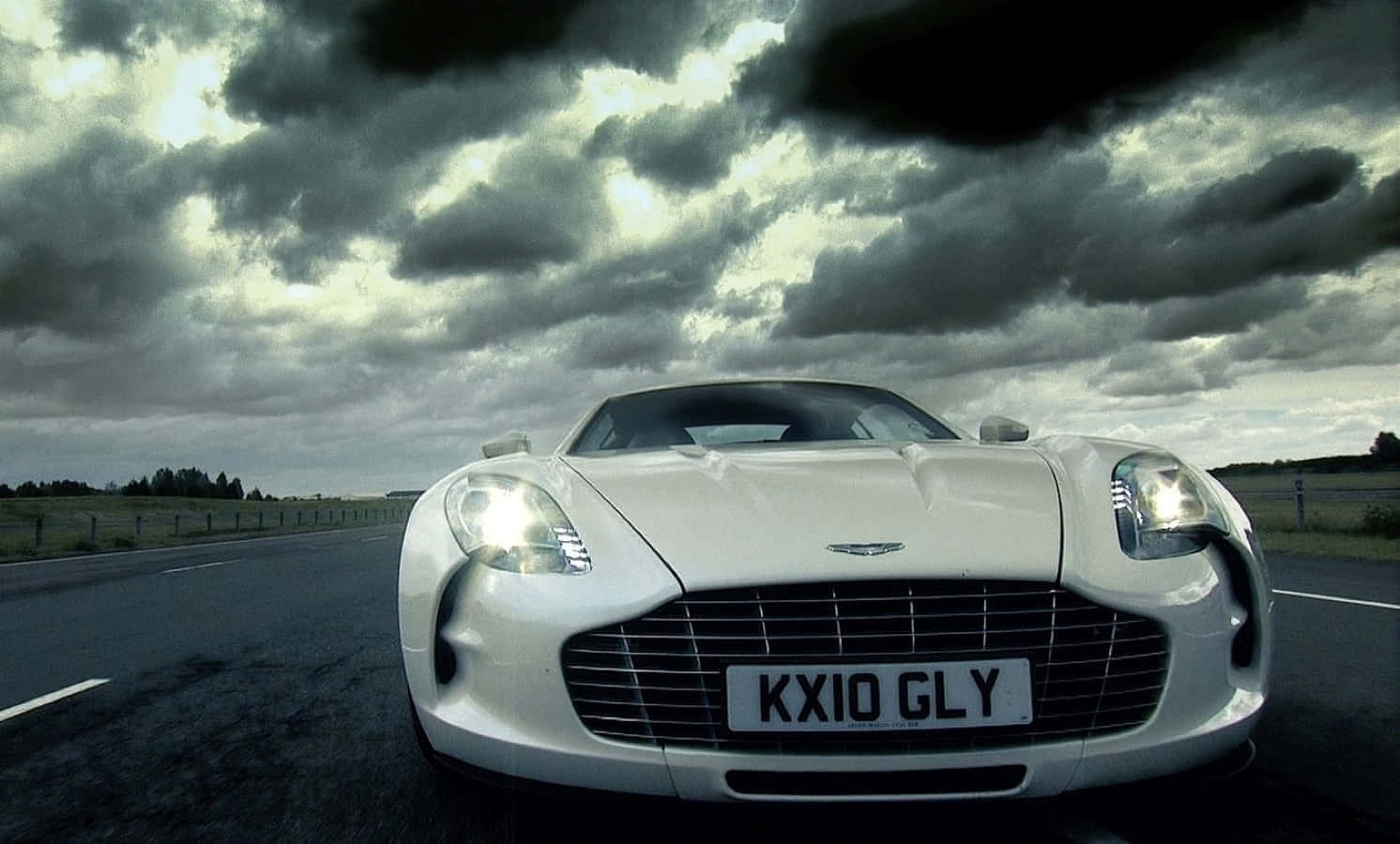 Aston Martin One-77 桌布 壁紙 — 一輛白色Aston Martin跑車亮著頭燈，在戲劇性的多雲天空下，於開闊的道路上面對鏡頭。