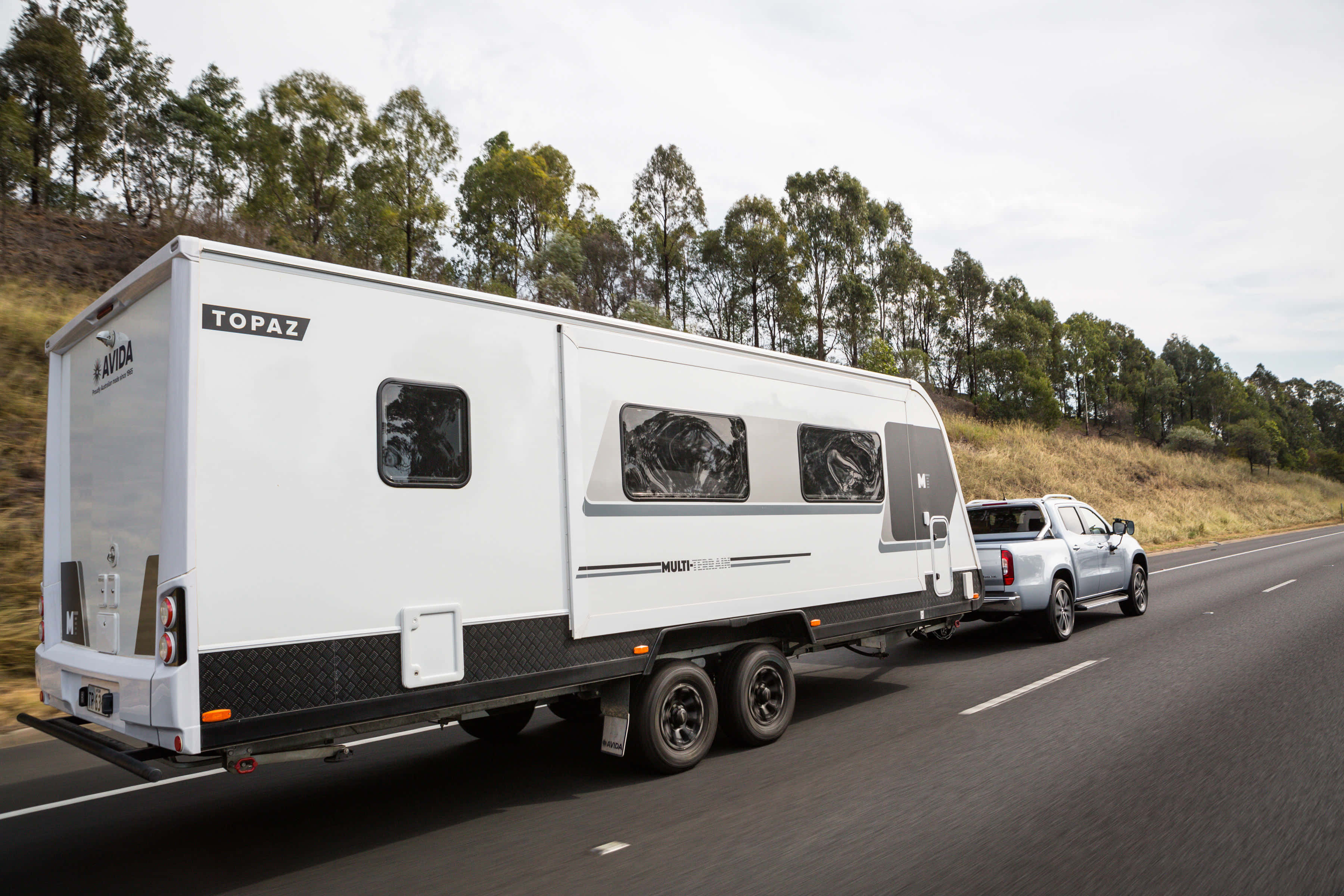 Caravan 桌布 壁紙 — 一輛白色Avida Topaz露營車正被一輛銀色小型貨車拖行，在鋪砌的道路上穿過樹木繁茂的山坡。