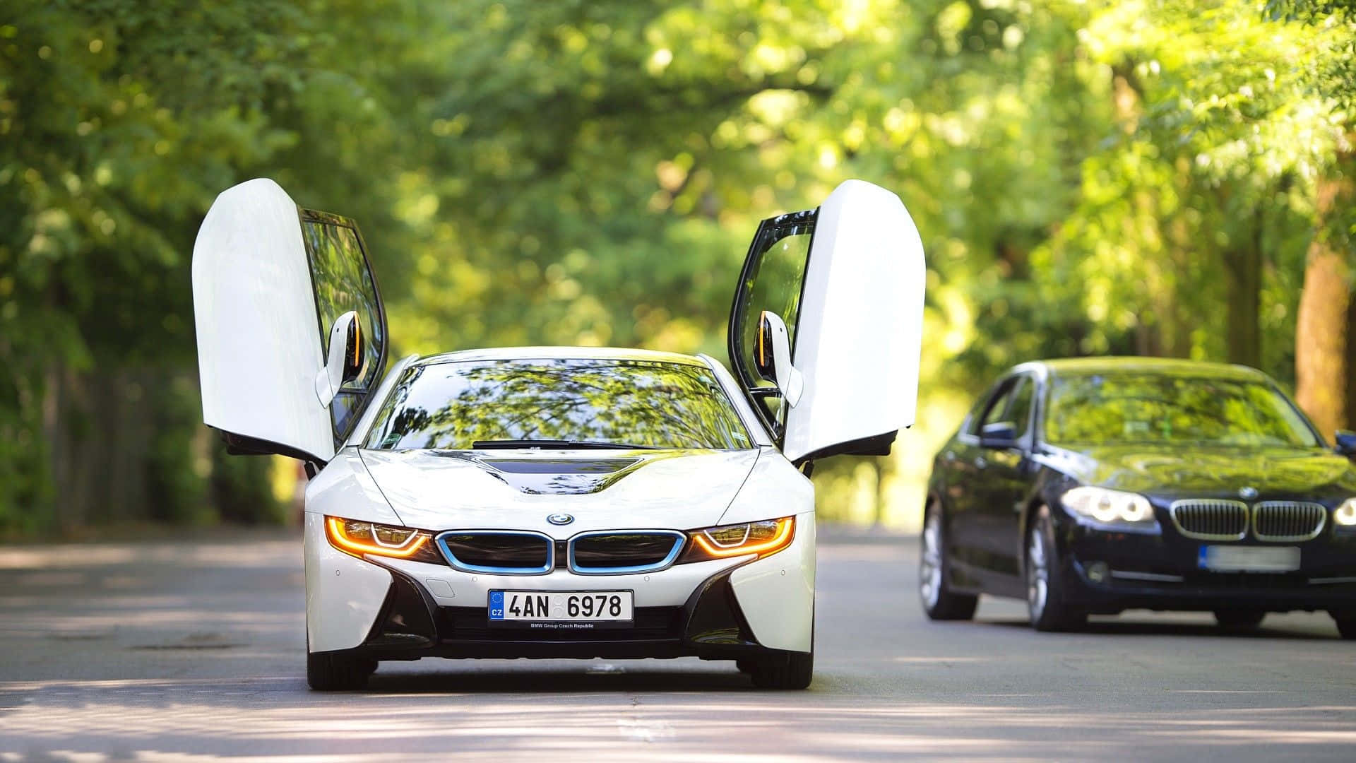 BMW 動圖 桌布 壁紙 — 一輛白色BMW i8打開鷗翼車門，在陽光明媚的林蔭大道上滑行，後面跟著一輛黑色BMW轎車。