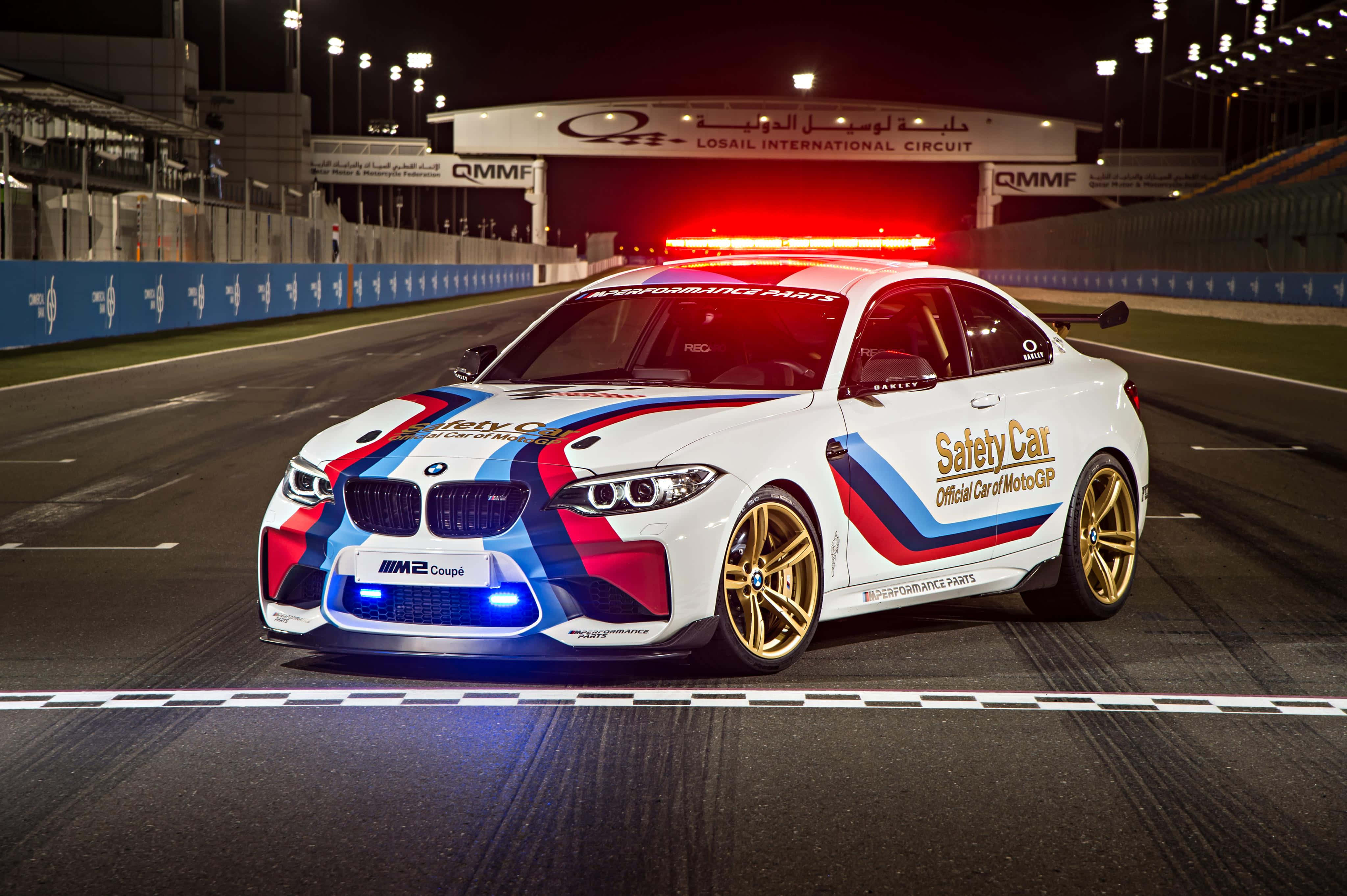 Safety Car 桌布 壁紙 — 一輛白色BMW M2 Coupe安全車，帶有紅、藍、金色塗裝，在夜間有照明的MotoGP賽道上奔馳。