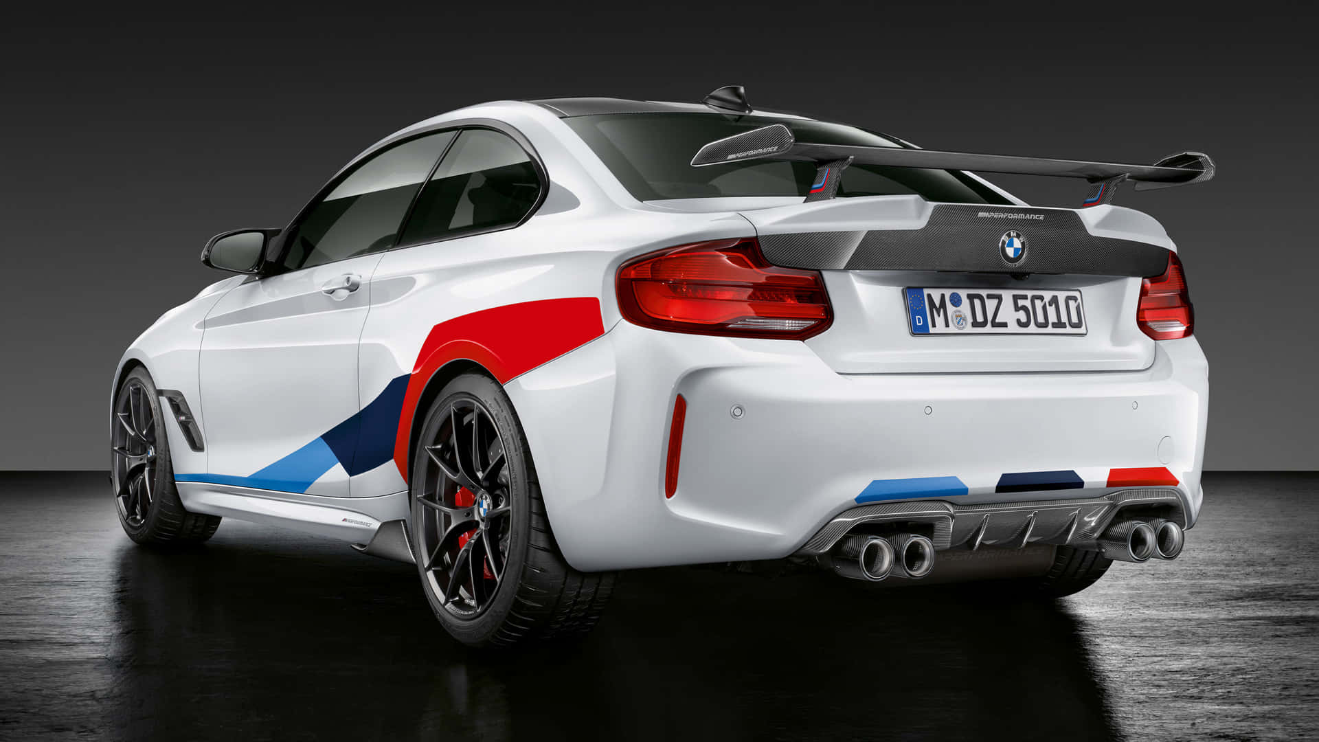 BMW 動圖 桌布 壁紙 — 一輛白色BMW M2雙門跑車，帶有紅藍賽車條紋和大型尾翼，在黑暗的攝影棚中以車尾四分之三角度展示。