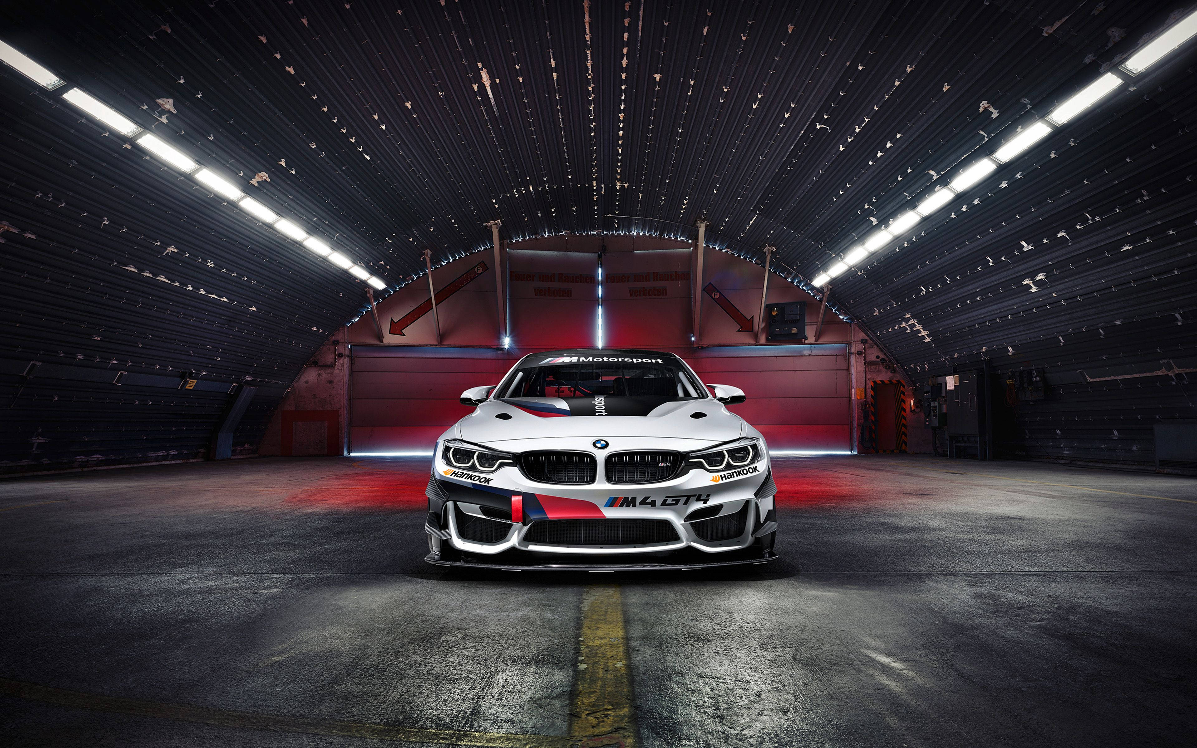 BMW 動圖 桌布 壁紙 — 一輛白色BMW M4 GT4賽車帶有紅色點綴，在黑暗的工業車庫中，明亮的頂燈下居中停放。