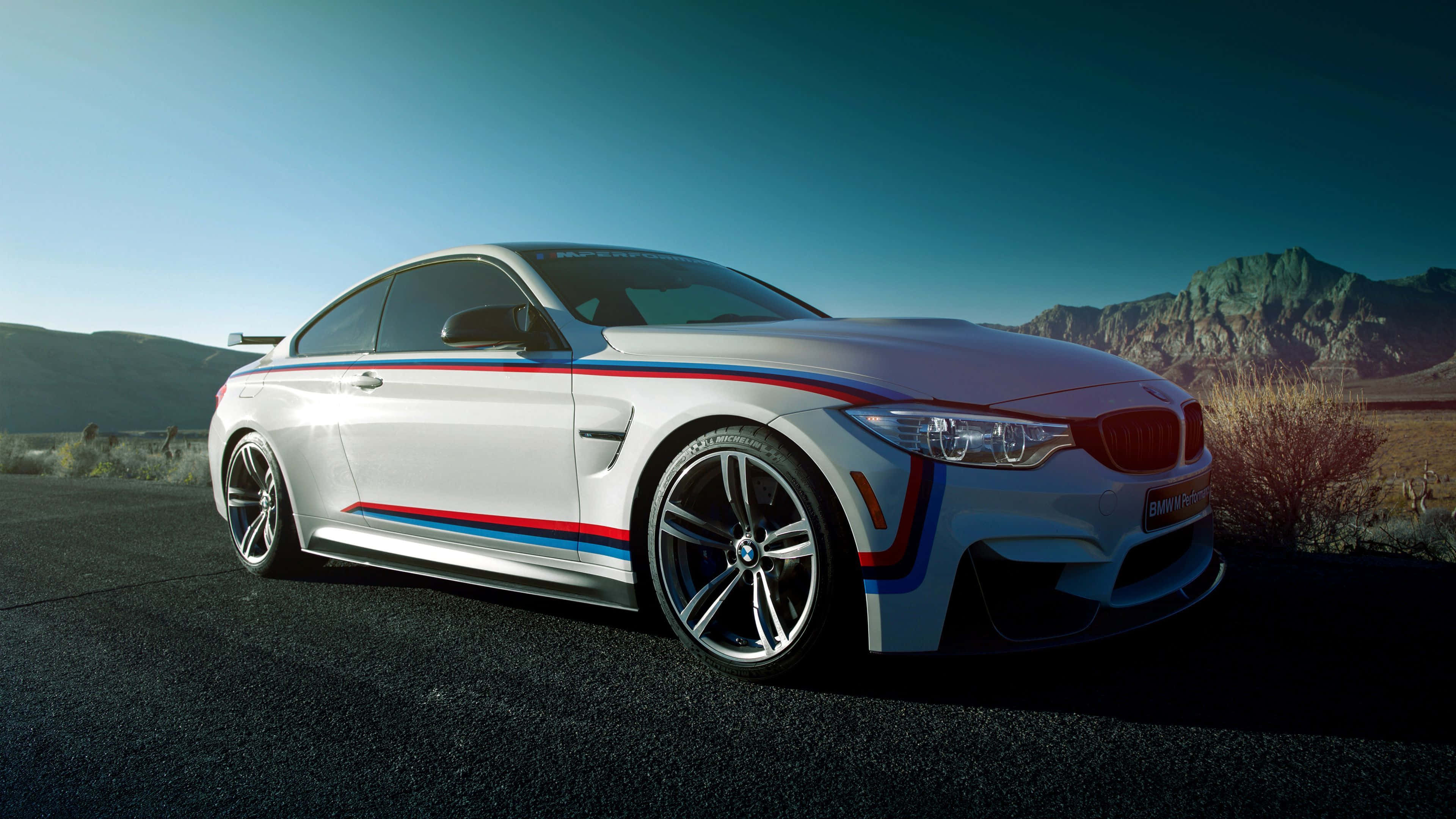跑車 桌布 壁紙 — 一輛白色BMW M4，帶有藍色、紅色和白色的賽車條紋，停在沙漠公路上，背景是山脈，天空清澈湛藍。