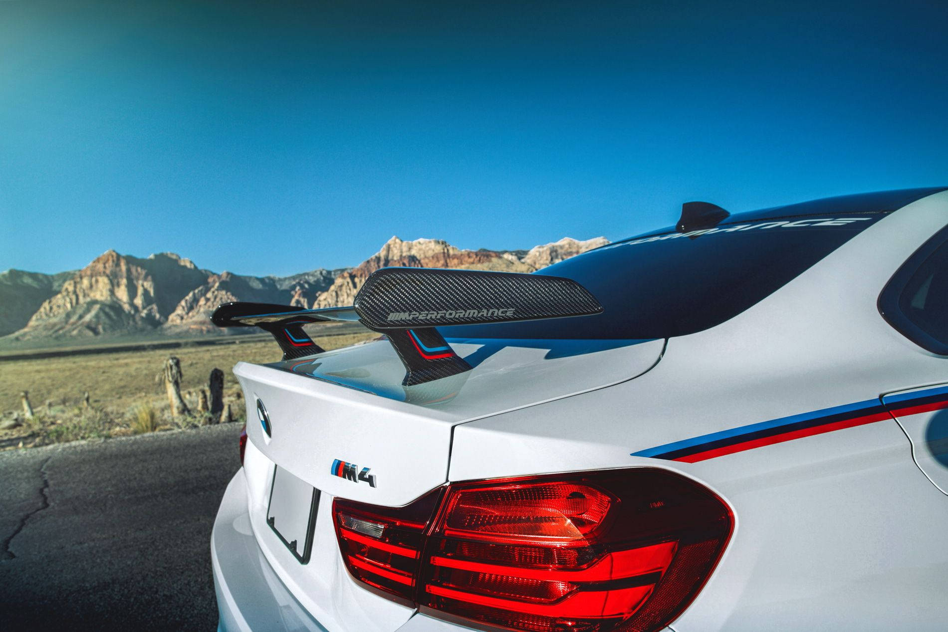 m 桌布 壁紙 — 一輛白色BMW M4配備碳纖維尾翼，停泊在沙漠道路上，背景是岩石山脈和清澈的藍天。