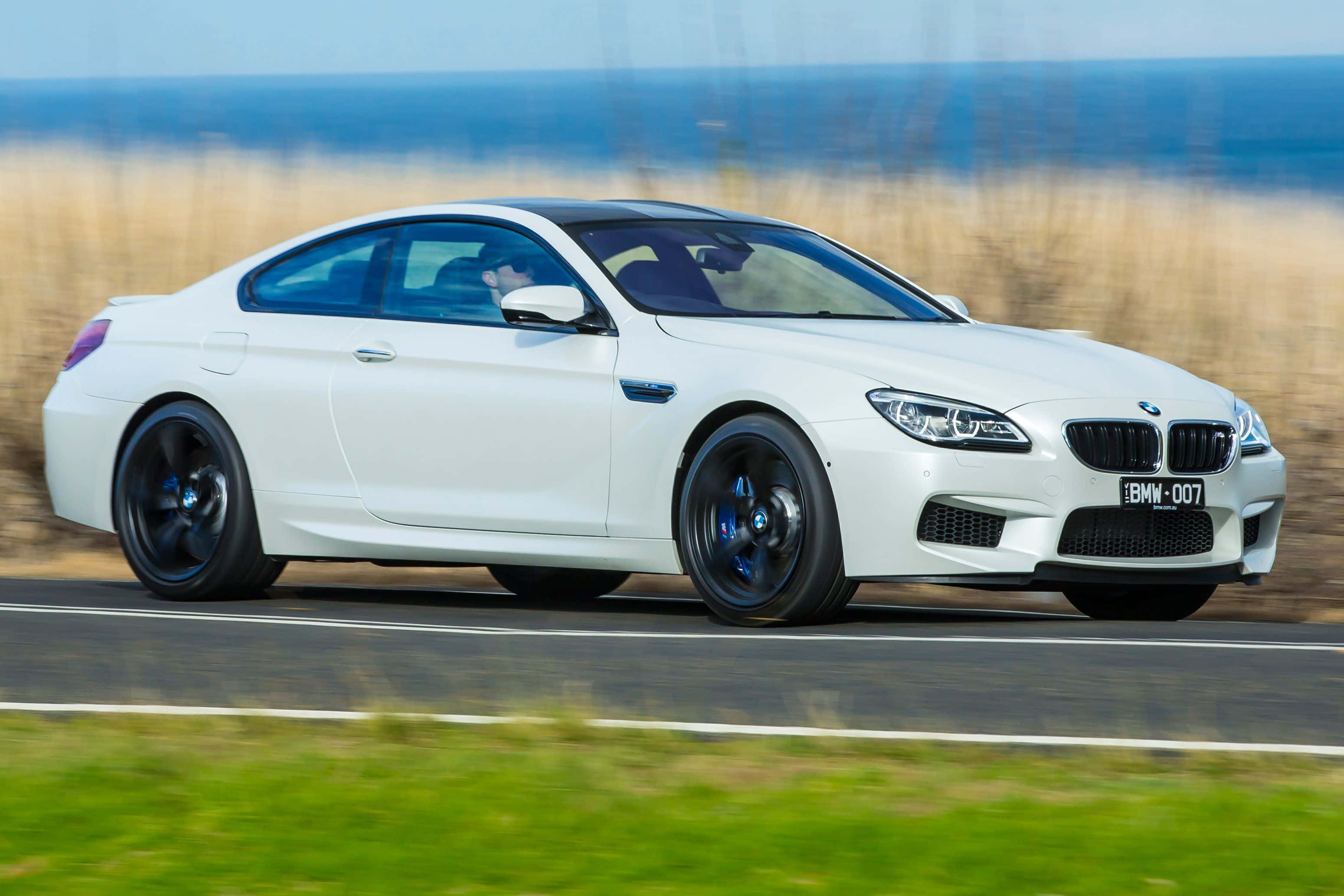 Bmw 6 Series 桌布 壁紙 — 一輛白色BMW M6雙門跑車沿著海岸公路疾駛，背景是模糊的草地和海洋。
