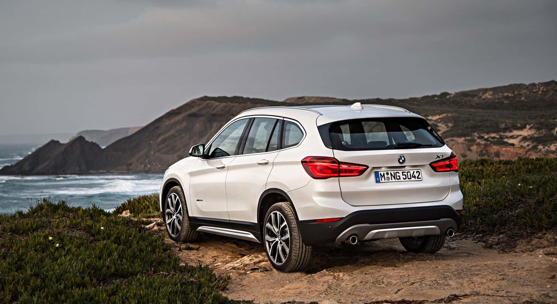 Bmw X1 桌布 壁紙 — 一輛白色BMW X1 SUV停泊在崎嶇的海岸懸崖上，俯瞰著下方波濤洶湧的海洋。