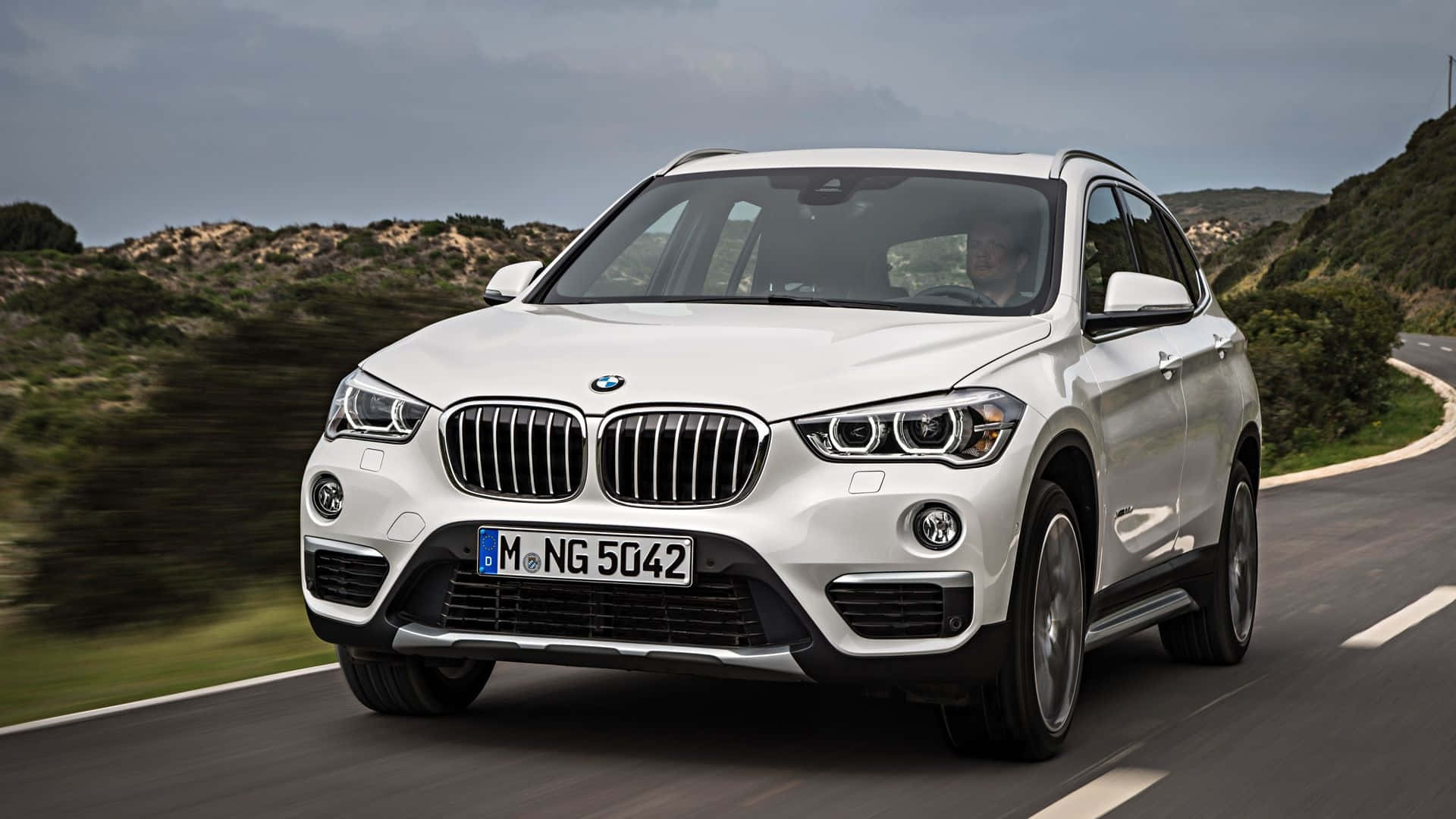 Bmw X1 桌布 壁紙 — 一輛白色BMW X1 SUV在陰天之下，沿著蜿蜒的道路疾馳，穿越丘陵起伏的綠色景觀。