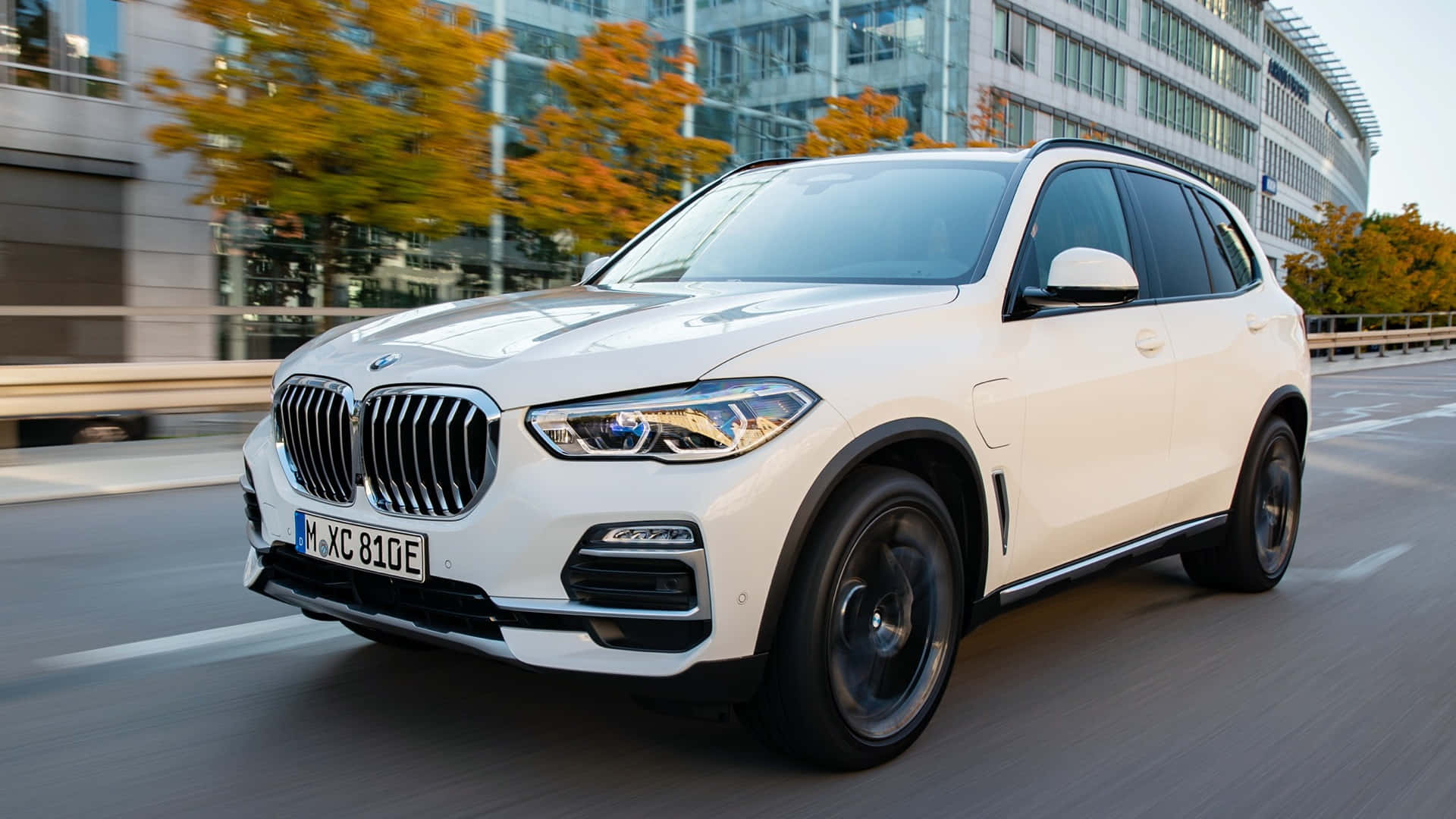 Bmw X5 桌布 壁紙 — 一輛白色BMW X5 xDrive45e SUV在城市的街道上行駛，經過秋天的樹木和玻璃建築。