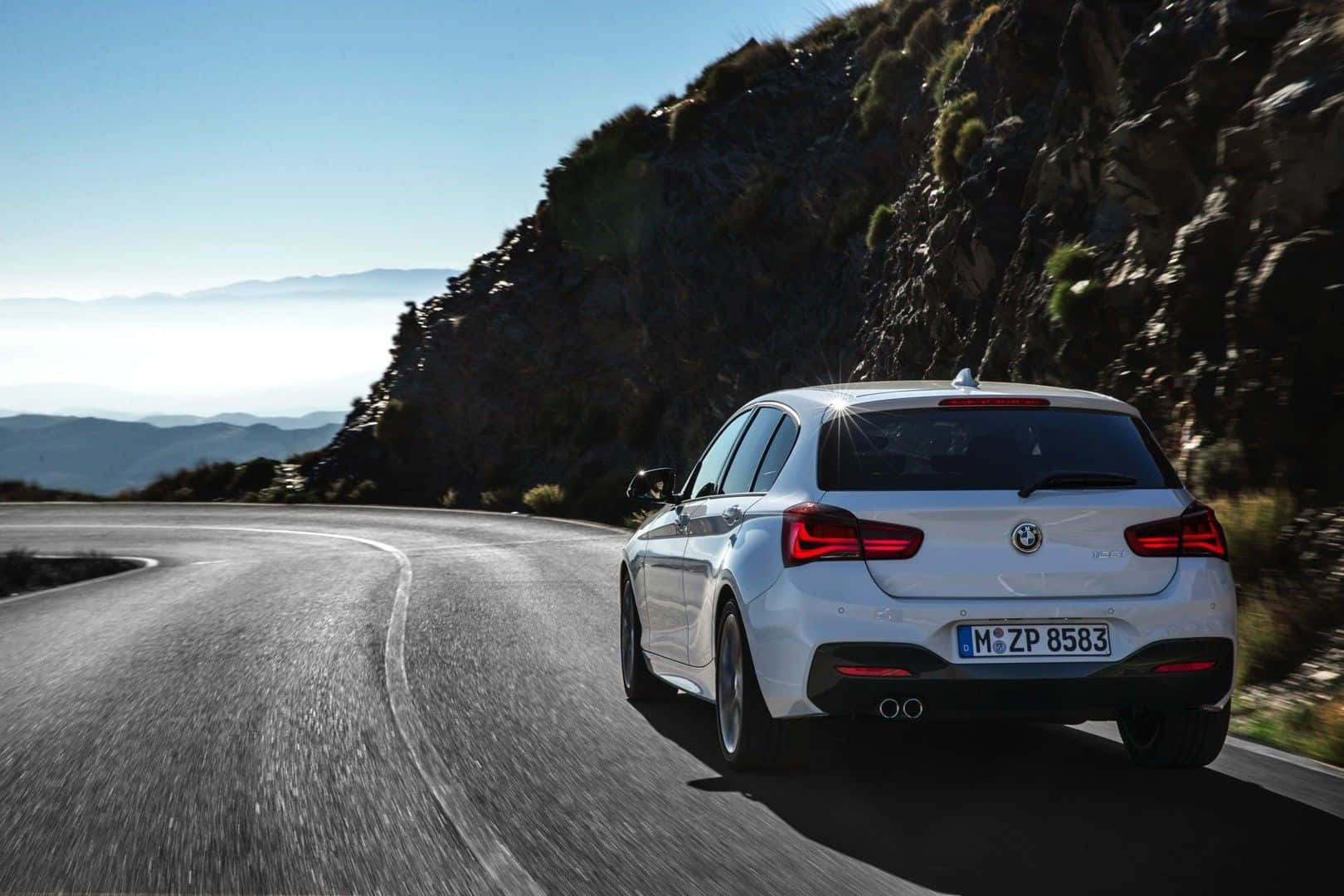 Bmw 1 Series 桌布 壁紙 — 一輛白色BMW掀背車沿著蜿蜒的山路行駛，兩旁是岩石峭壁和遠處的山丘，天空清澈湛藍。