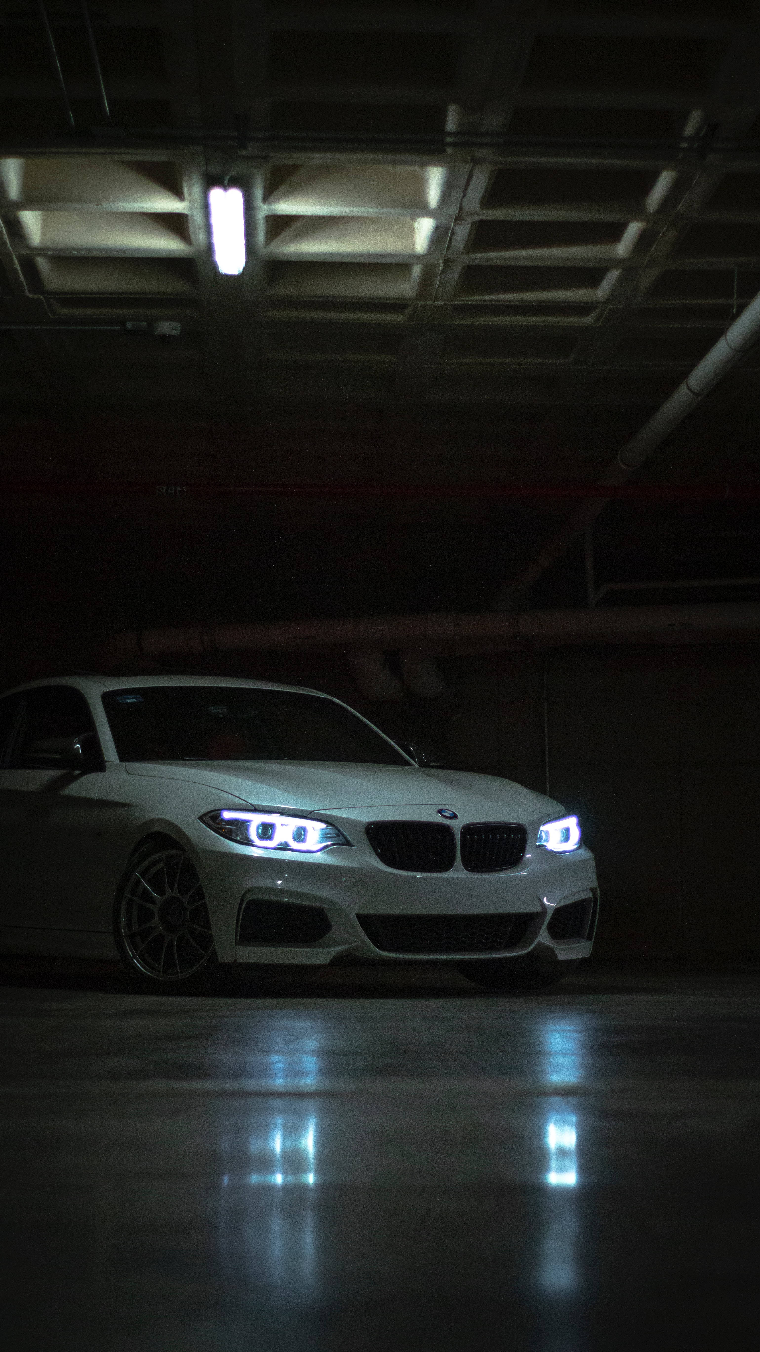 Bmw M Series 桌布 壁紙 — 一輛白色BMW跑車，車頭燈閃爍，停放在燈光昏暗的地下停車場，在濕漉漉的混凝土地板上投射出清晰的倒影。