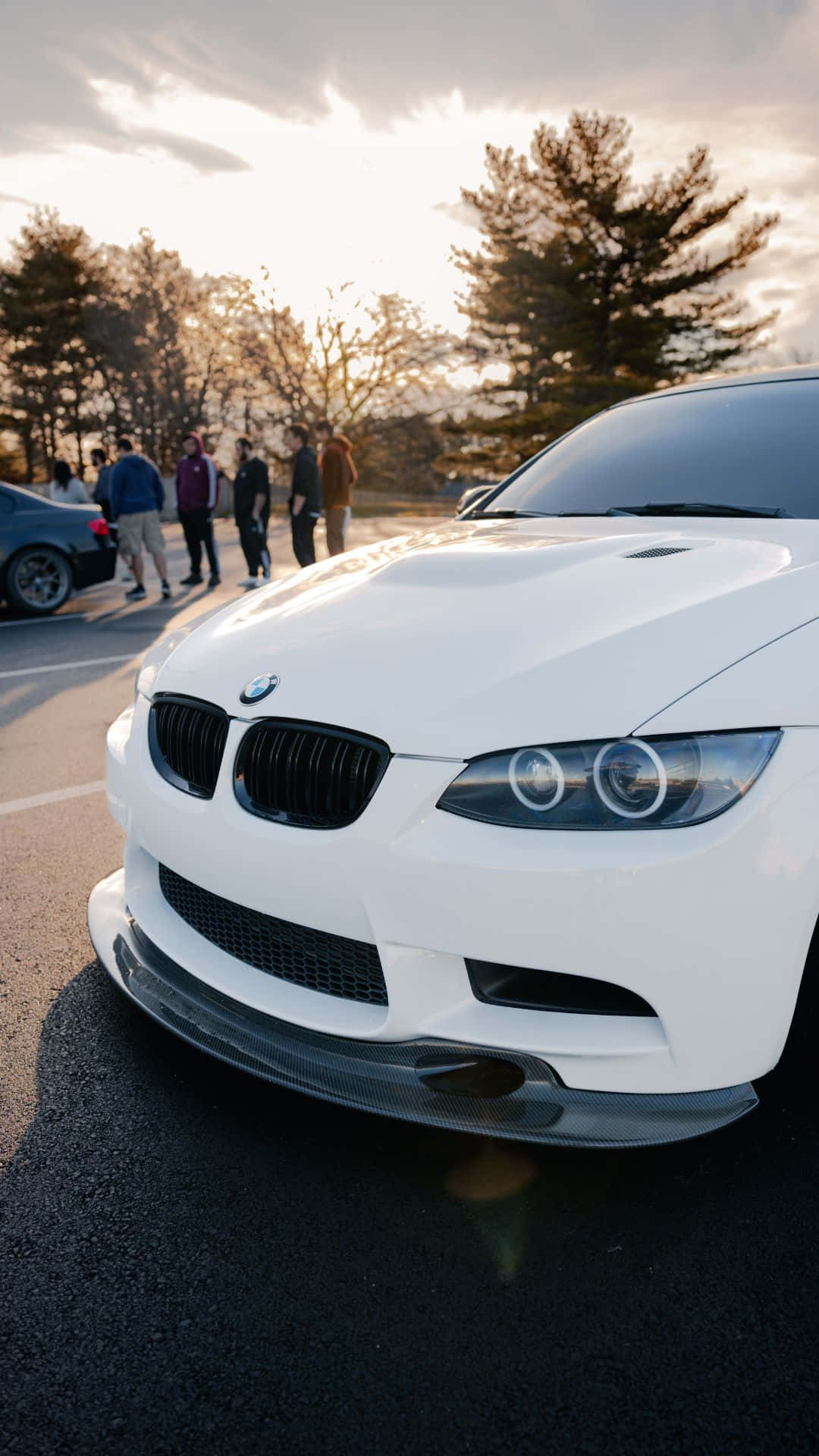 Android Bmw 桌布 壁紙 — 一輛白色BMW跑車在金色夕陽下閃閃發光，停車場附近有人群聚集。
