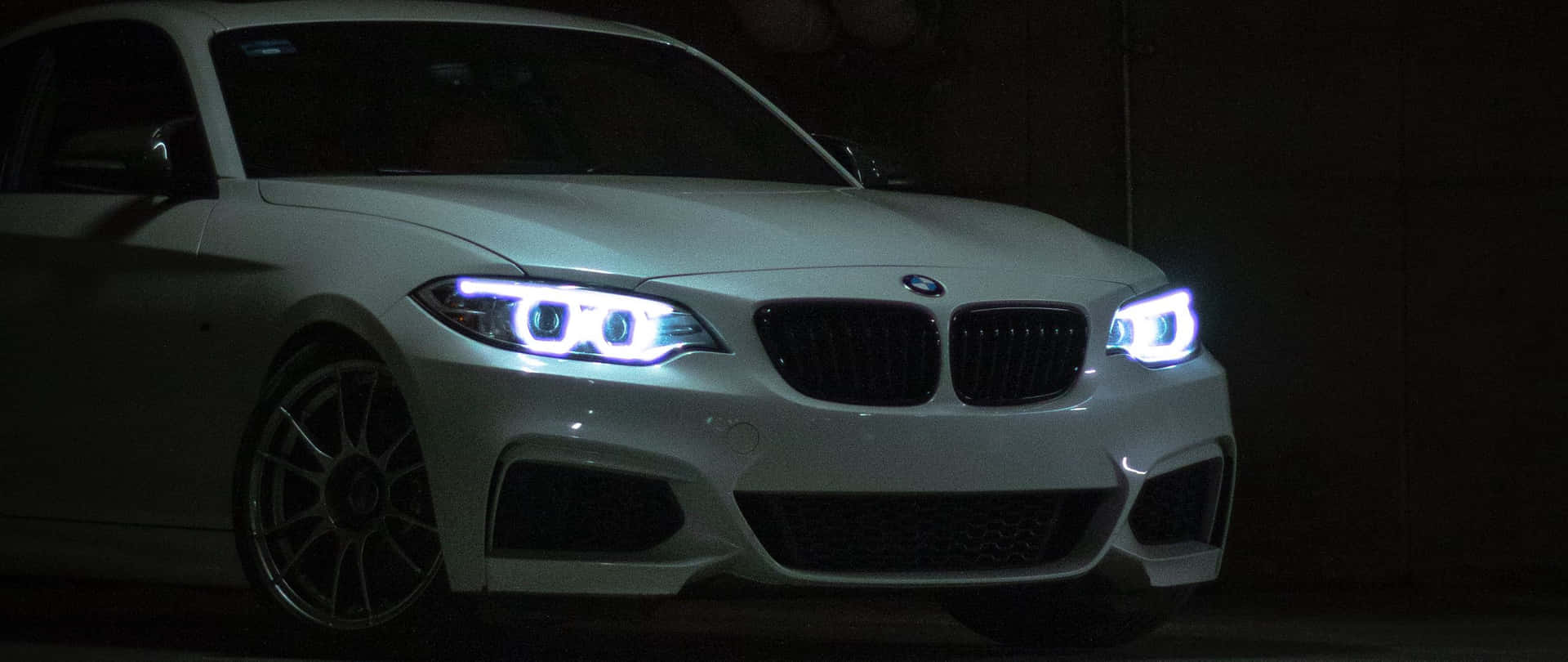 BMW 動圖 桌布 壁紙 — 一輛白色BMW跑車在黑暗的車庫中發出明亮的藍色車頭燈，在其流線型車身上投下銳利的陰影。