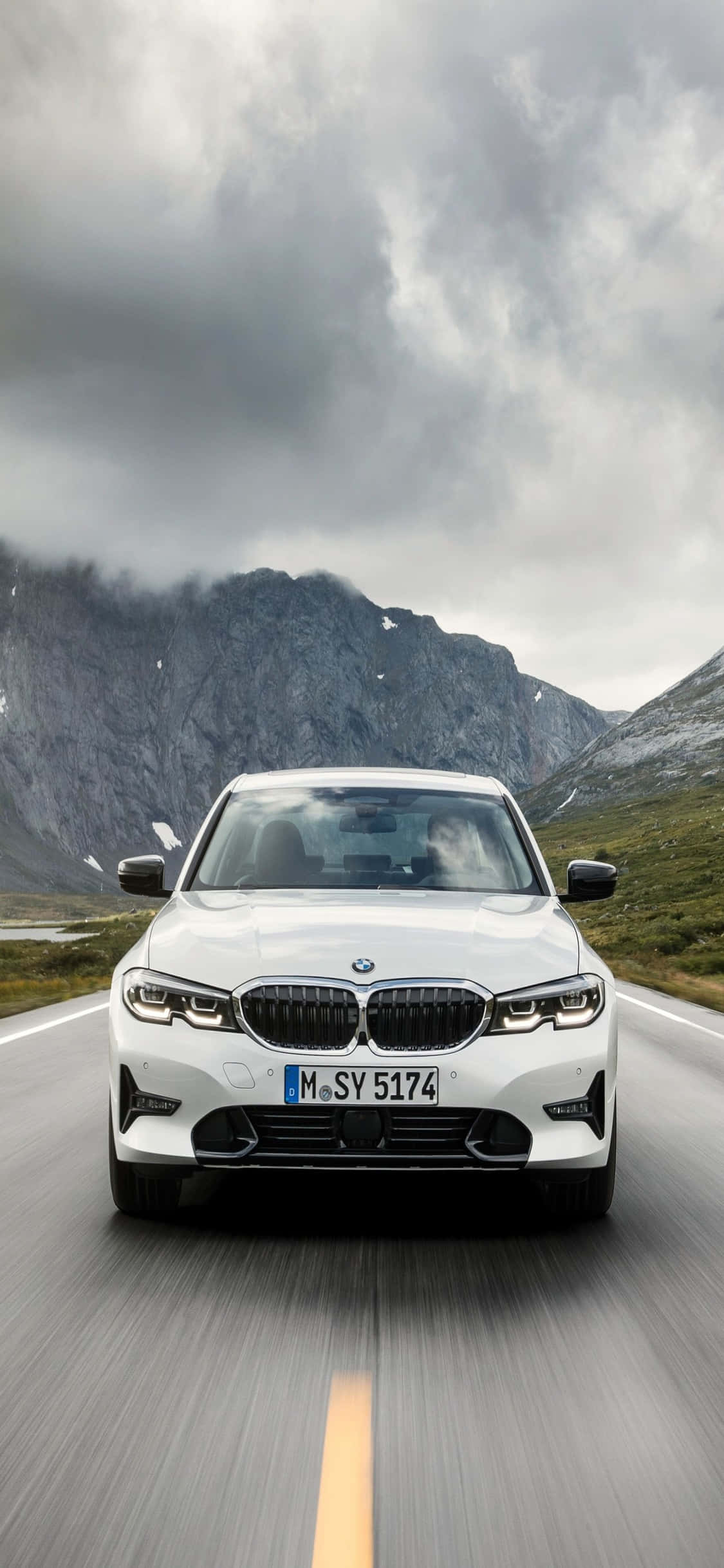 iPhone X Bmw 桌布 壁紙 — 一輛白色BMW轎車在戲劇性的多雲天空下，背景是崎嶇的山峰，沿著山路疾馳。