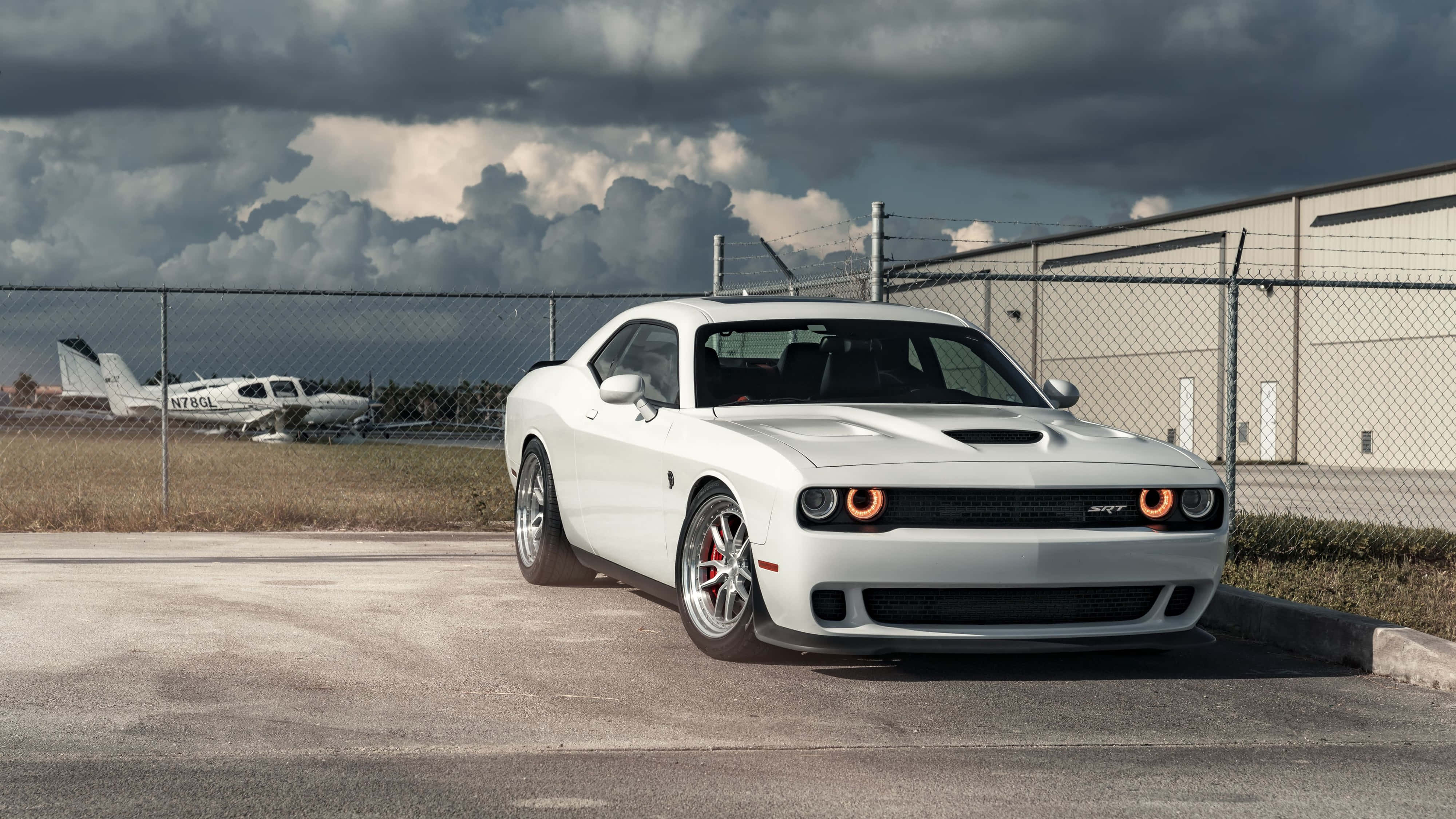 Dodge 桌布 壁紙 — 一輛白色Dodge Challenger SRT停放在瀝青路上，靠近鐵絲網圍欄，後面有一架小型飛機，在戲劇性的多雲天空下可見。