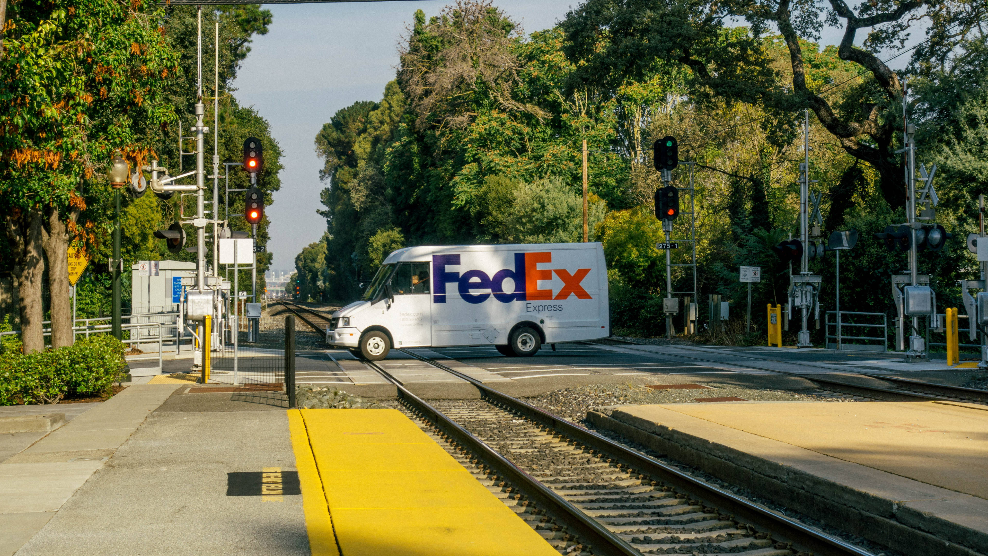 聯邦快遞 桌布 壁紙 — 一輛白色FedEx Express貨車在明亮的紅色警告燈下停於鐵路平交道，周圍是茂密的綠樹。