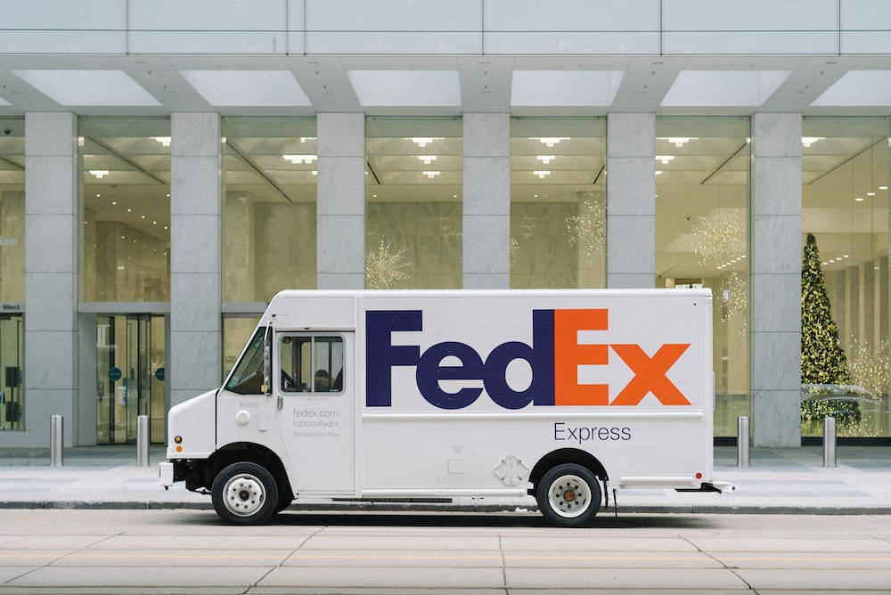 聯邦快遞 桌布 壁紙 — 一輛白色FedEx Express送貨車停在一棟現代玻璃帷幕建築外，建築內可見一棵亮燈的聖誕樹。