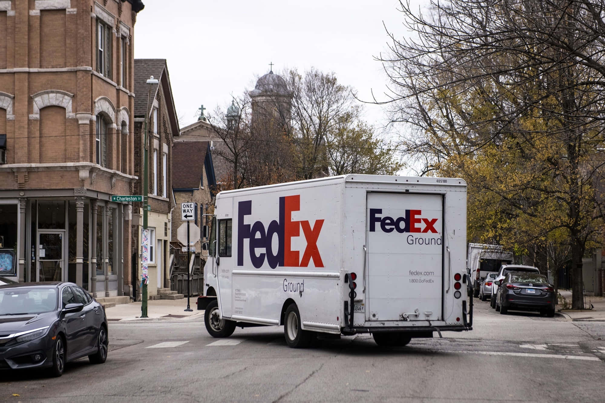 聯邦快遞 桌布 壁紙 — 一輛白色FedEx Ground貨車在陰天下行駛在一條綠樹成蔭的城市街道上，經過磚砌建築和停泊的汽車。