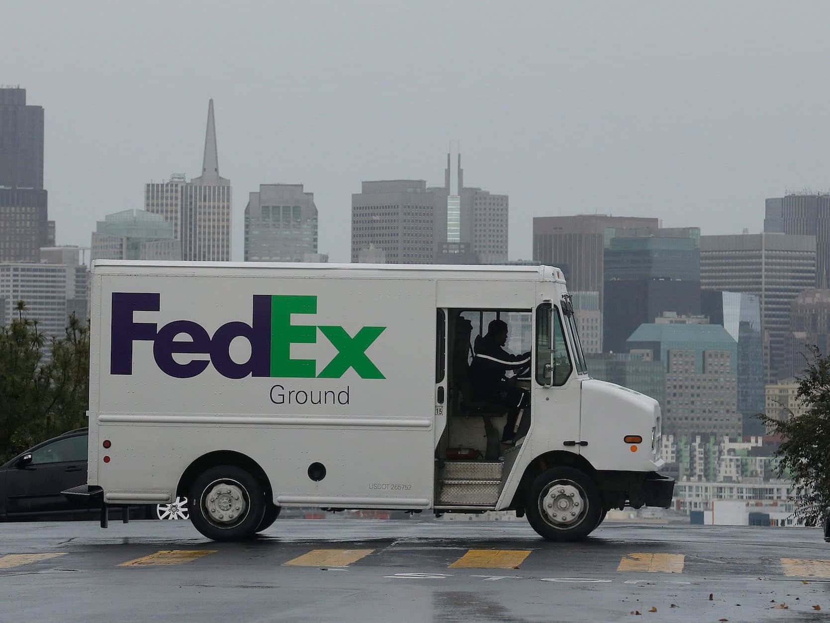 聯邦快遞 桌布 壁紙 — 一輛白色FedEx Ground貨車停放在潮濕的市區空地，背景是朦朧的城市天際線。