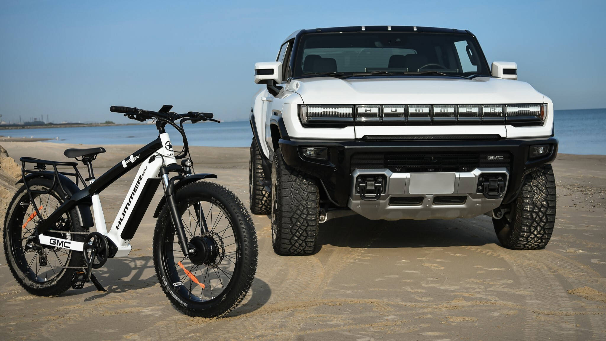 Truck 桌布 壁紙 — 一輛白色GMC Hummer EV SUV和匹配的電動胖胎自行車並排停放在沙灘上，背景是海洋和晴朗的天空。