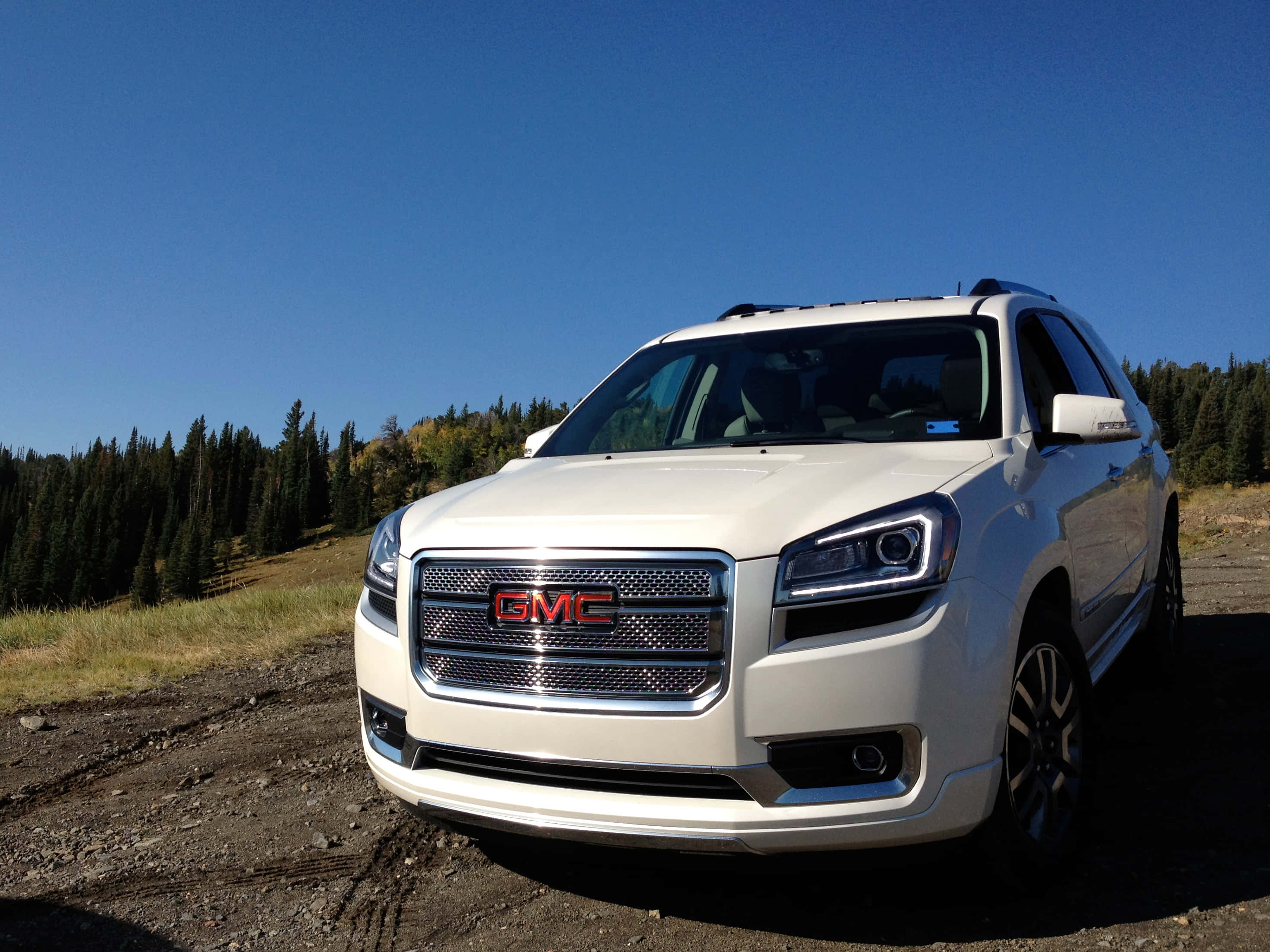 Gmc Acadia 桌布 壁紙 — 一輛白色GMC SUV停泊在碎石路上，背景有松樹和晴朗的藍天。