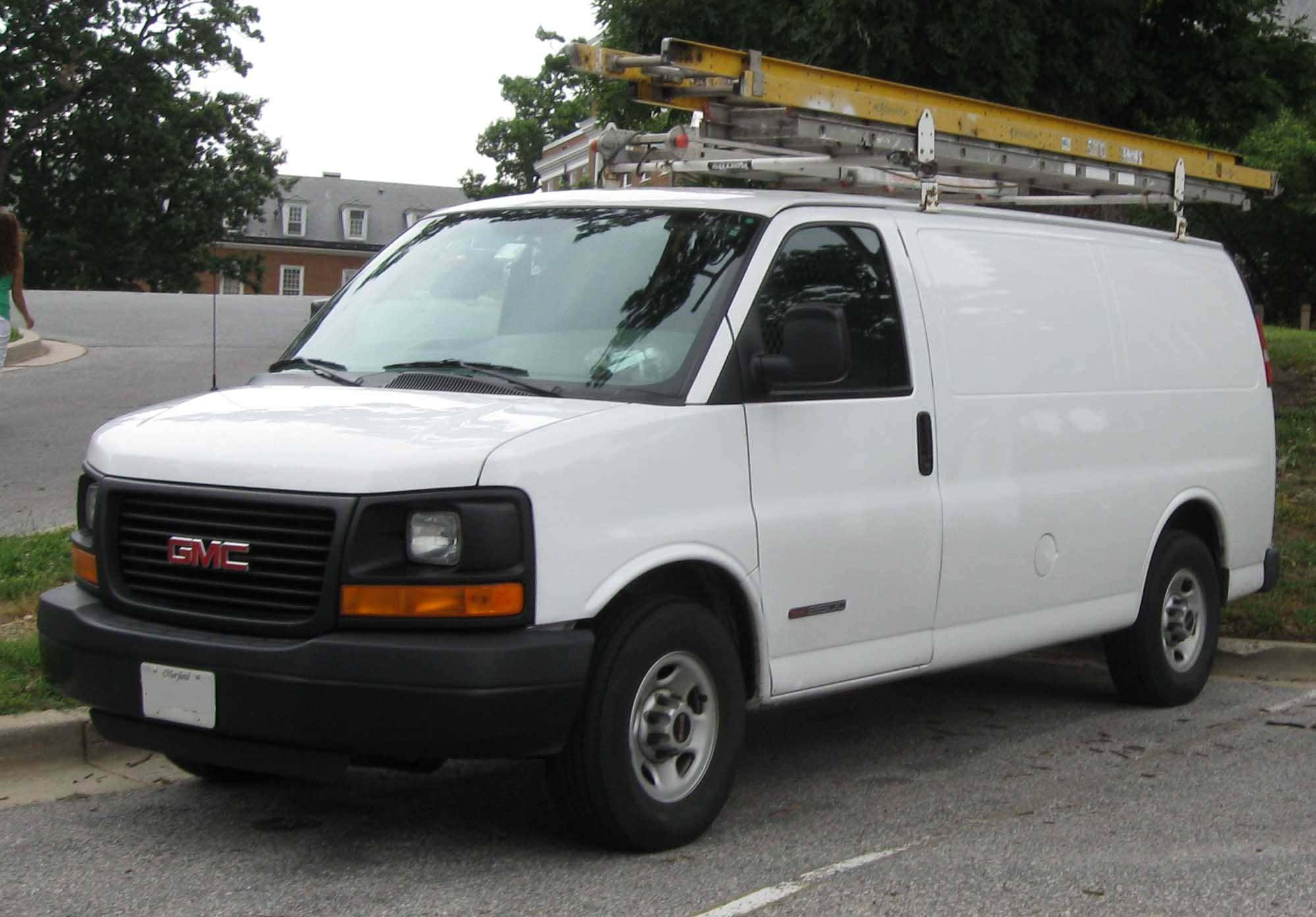 Gmc Savana 桌布 壁紙 — 一輛白色GMC廂型車，車頂裝有黃色梯子，停在路邊，旁邊有綠樹。