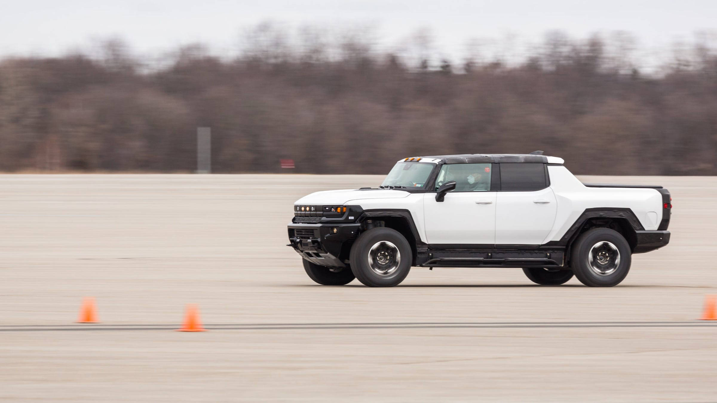 Truck 桌布 壁紙 — 一輛白色Hummer EV電動皮卡車在廣闊的混凝土測試跑道上繞著橙色錐體行駛，背景有模糊的樹木。