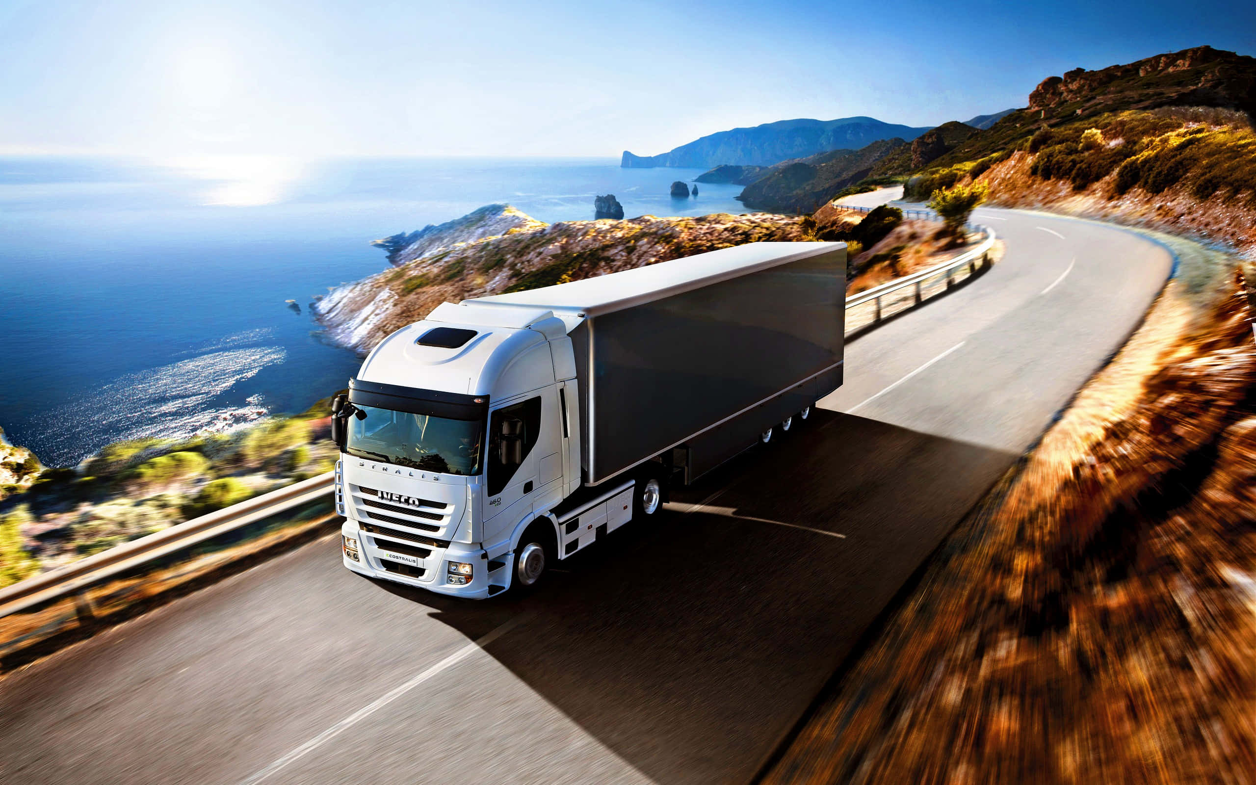 Semi Truck 桌布 壁紙 — 一輛白色IVECO半掛卡車沿著海岸高速公路疾馳，在明亮的藍色大海和陽光照耀的懸崖背景下呈現動態模糊效果。