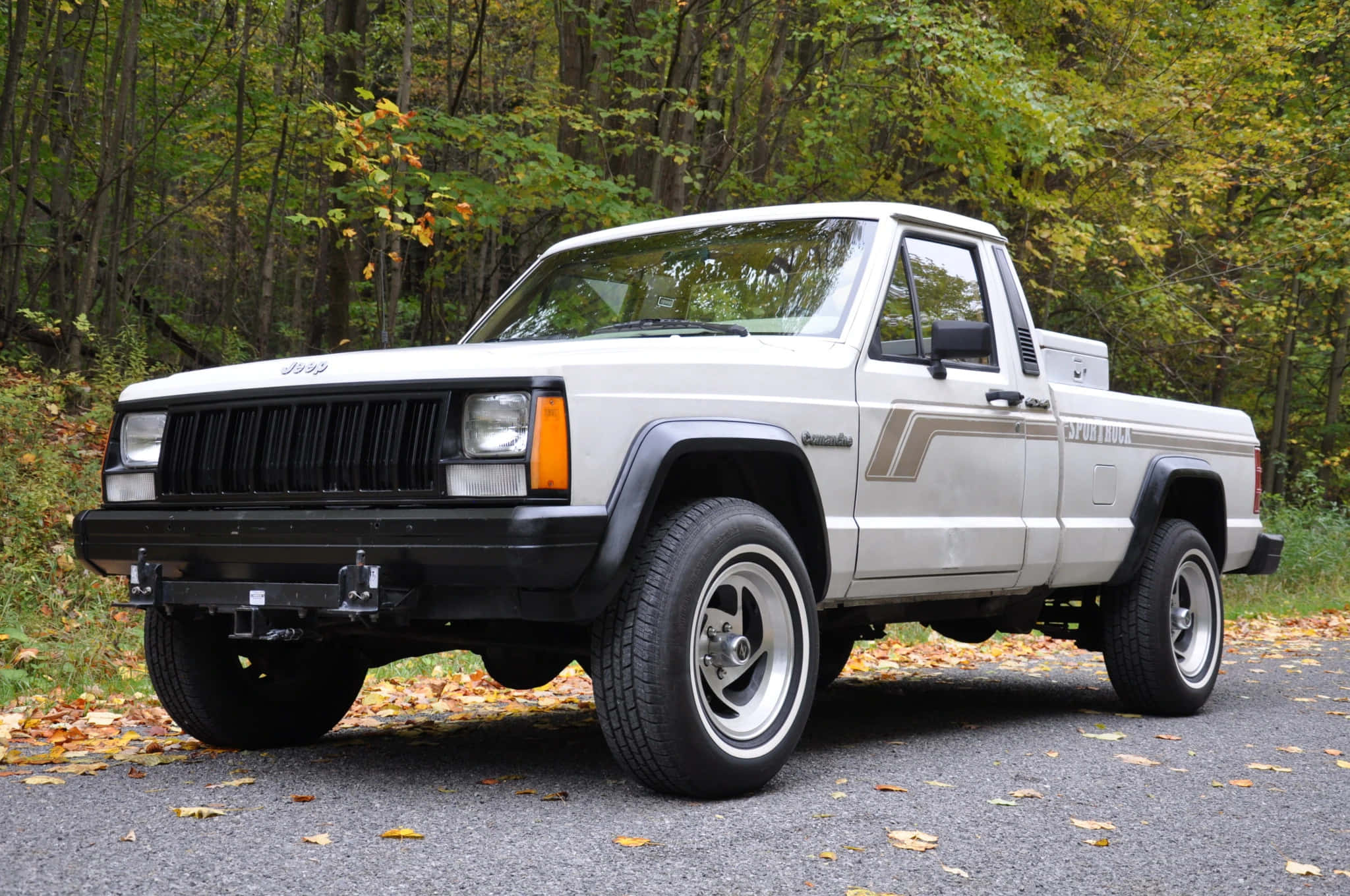 Jeep Comanche 桌布 壁紙 — 一輛白色Jeep Cherokee皮卡車停在碎石路上，周圍是秋日森林，落葉飄飄。