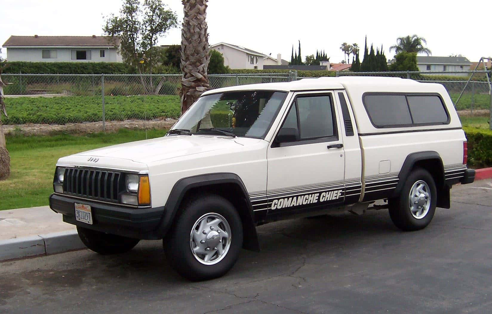 Jeep Comanche 桌布 壁紙 — 一輛白色Jeep Cherokee Comanche Chief旅行車停放在一個裂開的瀝青停車場，背景有棕櫚樹和郊區房屋。
