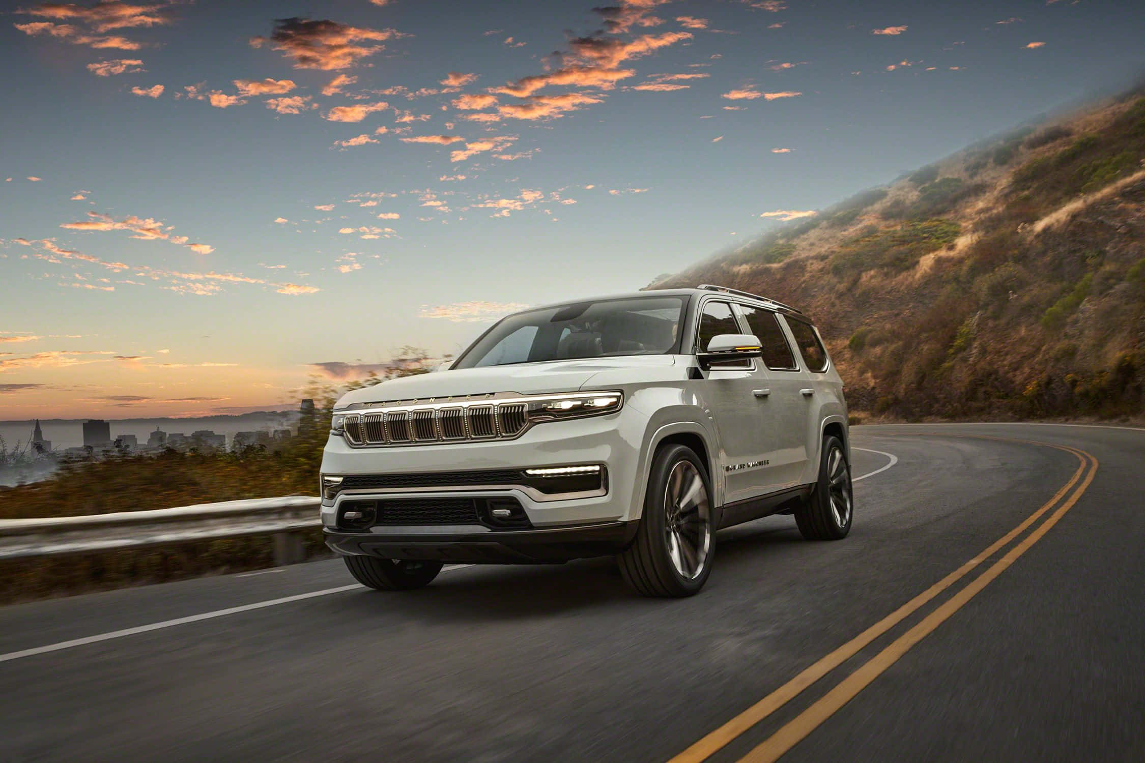 Jeep Wagoneer 桌布 壁紙 — 一輛白色Jeep Grand Wagoneer SUV在日落時分沿著蜿蜒的山路行駛，遠處是城市天際線。
